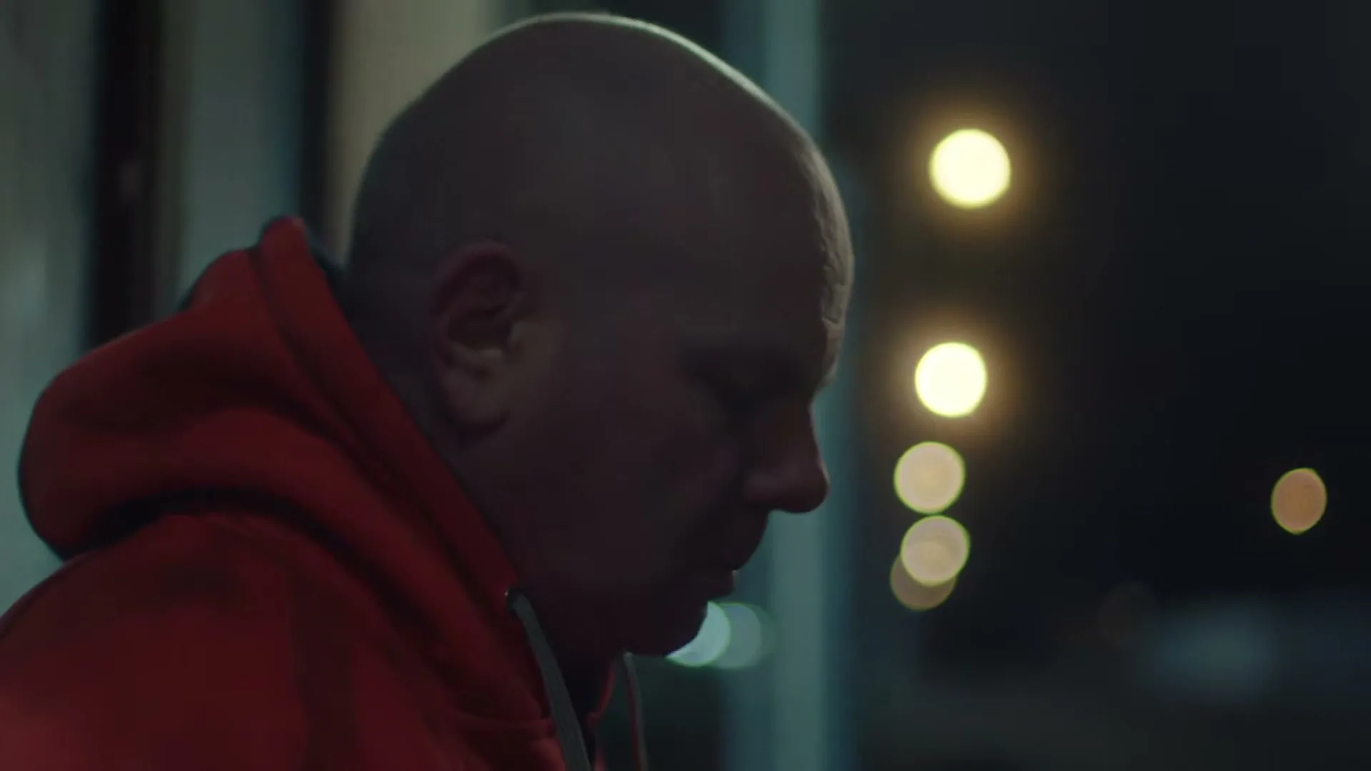 A bald man in a red hoodie looks down, appearing distressed, at night with blurred lights in the background.