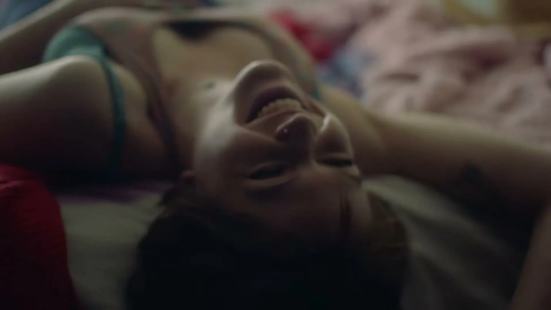 A young woman with short hair, wearing a tank top, lies on a bed, laughing joyfully with her head upside down.