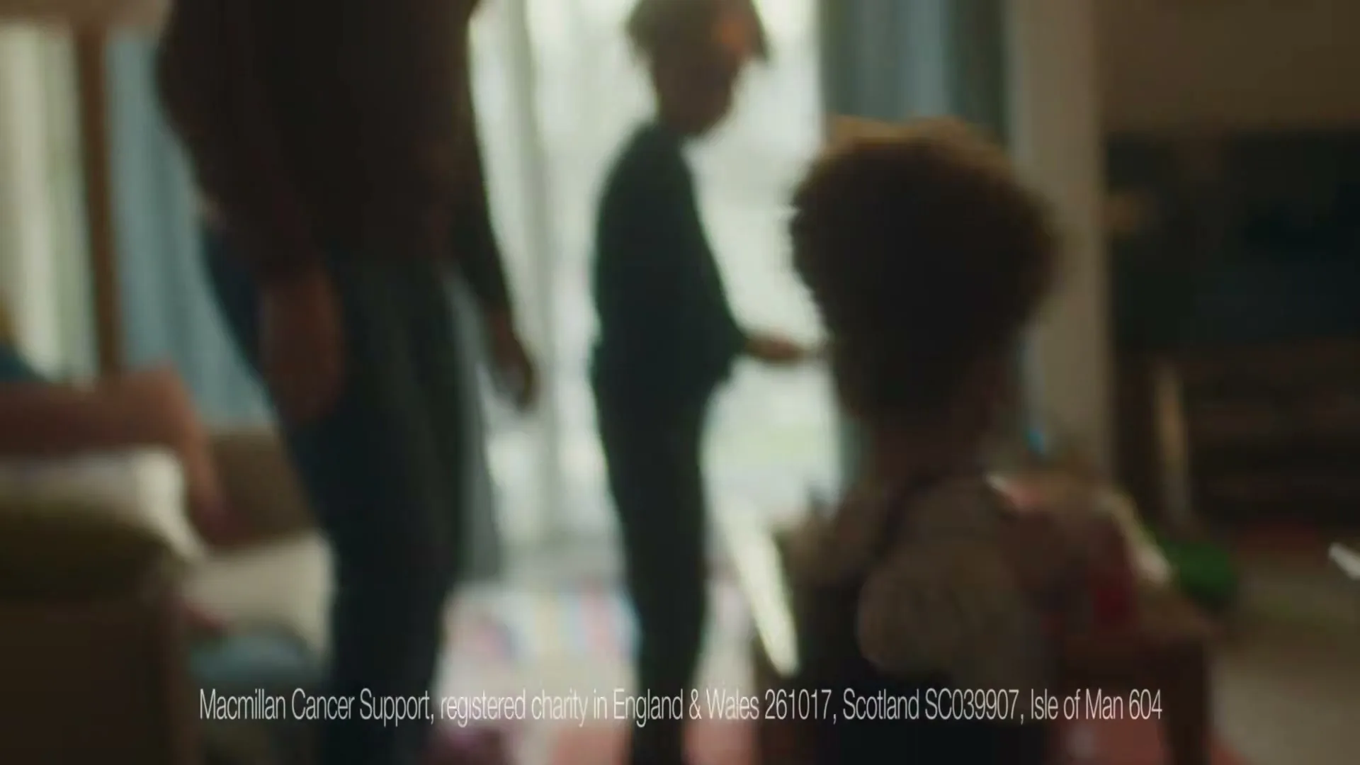A young child with curly hair walks away from the camera towards a man and another child in a softly lit living room.