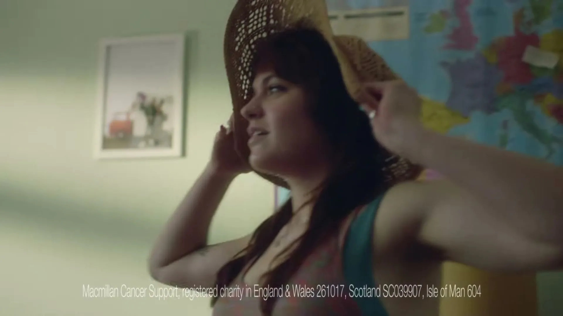 A young woman with dark hair tries on a straw hat in a bedroom with a map on the wall, looking happy and playful.