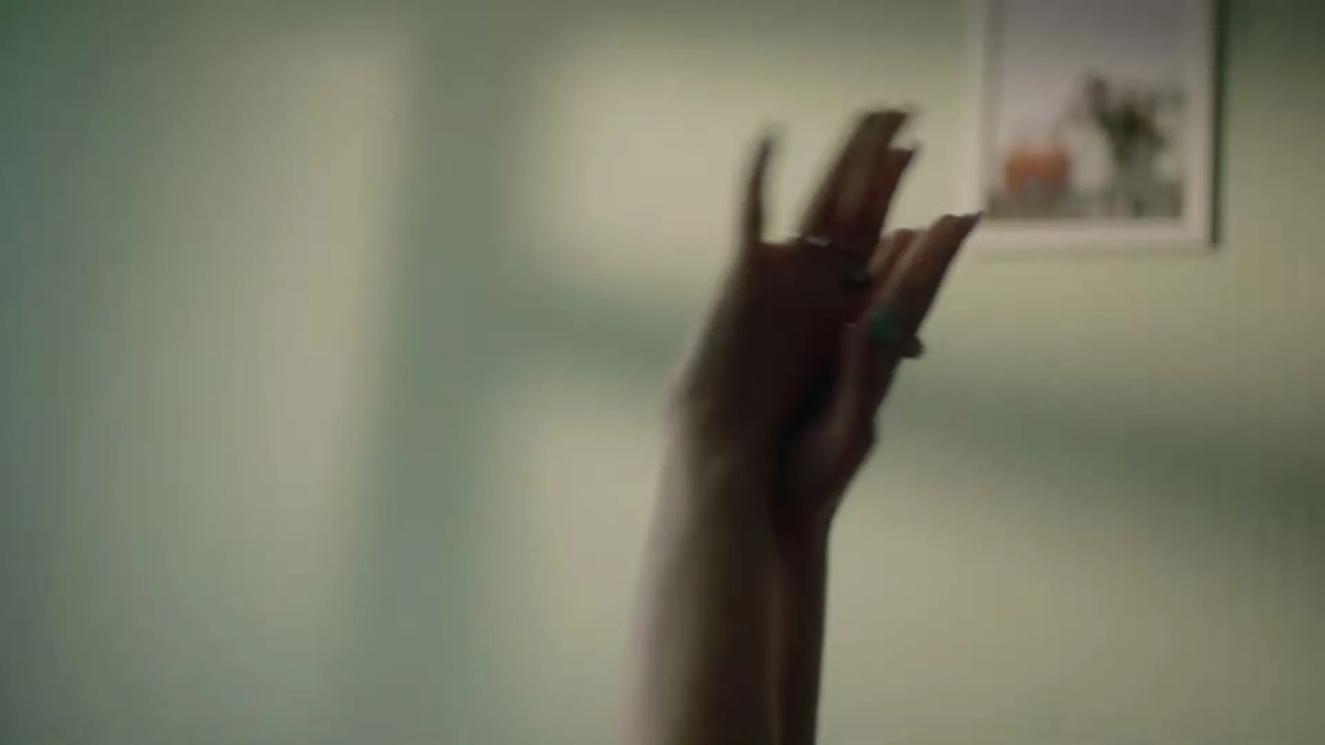 Two hands with rings and painted nails reach up and interlock against a light green wall with a framed picture.