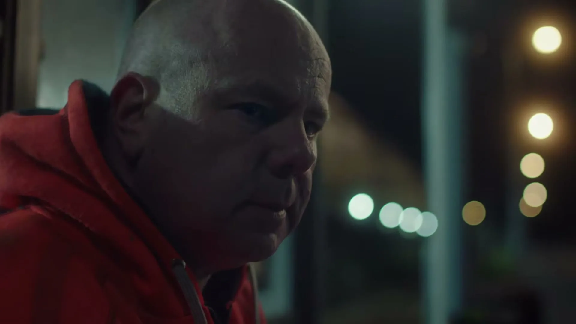 A bald man in his 50s, wearing a red hoodie, looks down with a worried expression at night.