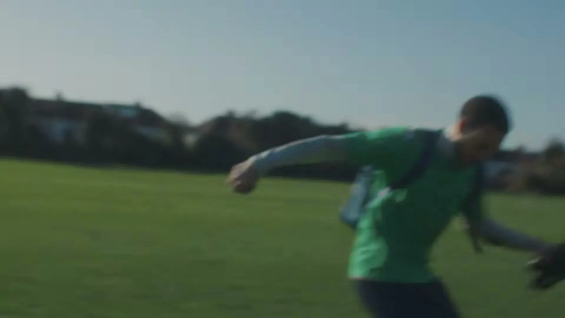 A young man in a green shirt and backpack runs across a sunny green field, holding something in his hand.