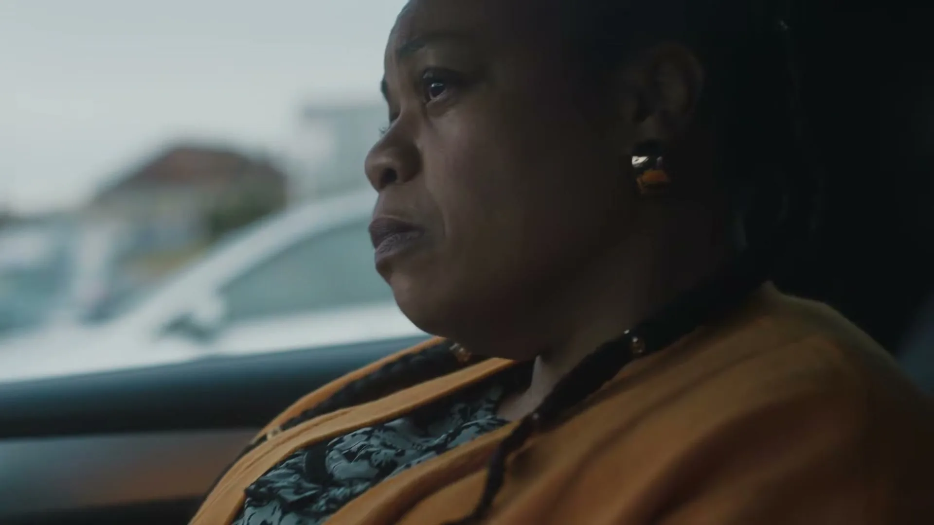 A woman in her 50s, wearing an orange cardigan, looks out the car window with a worried expression, while riding in a car.