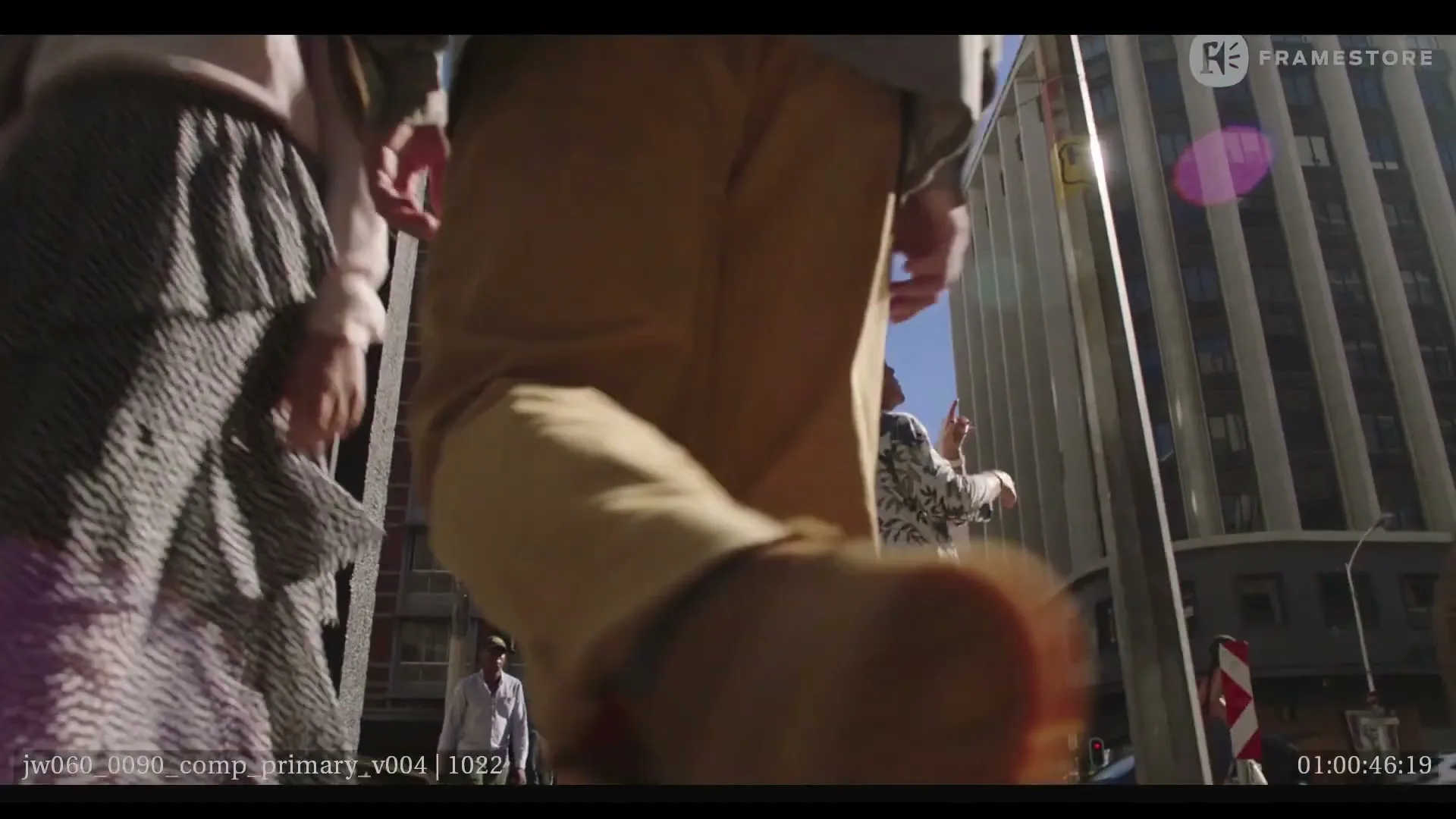 A low-angle shot of people walking on a sunny city street, with buildings in the background and a woman in orange walking towards the camera.