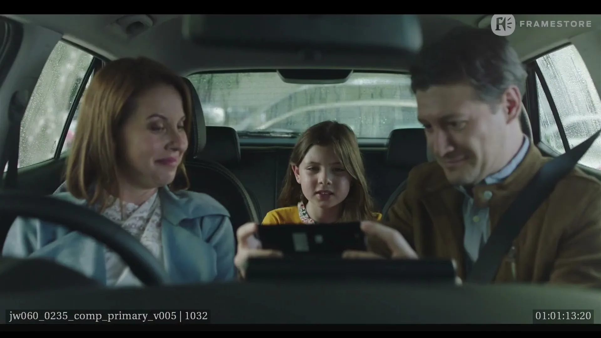 A family of three, a mother, father, and daughter, are sitting in a car, with the daughter holding a phone and the parents looking at her.