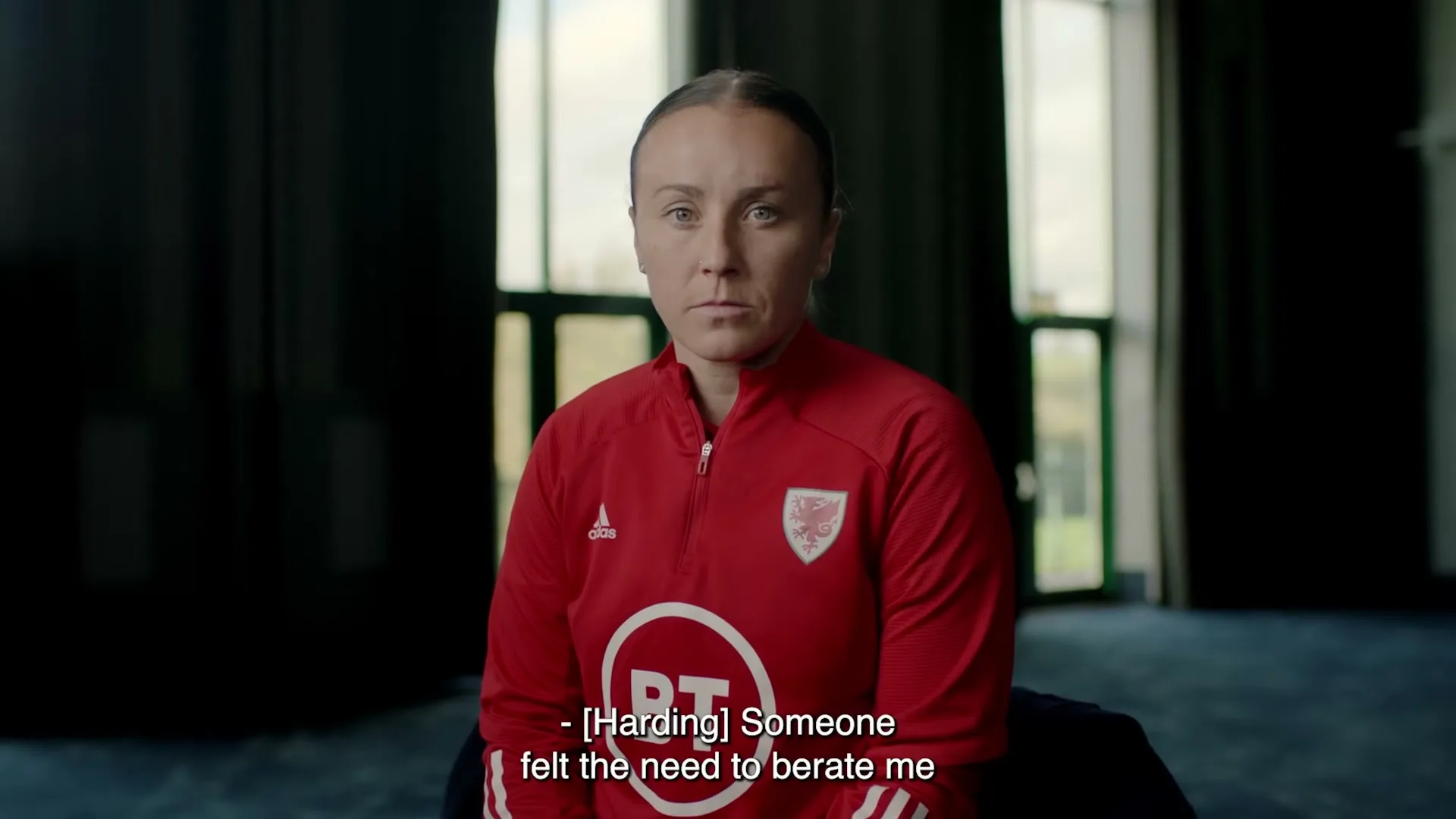 A female athlete in her 30s, wearing a red sports jacket, sits looking directly at the camera with a neutral expression in an interior setting.