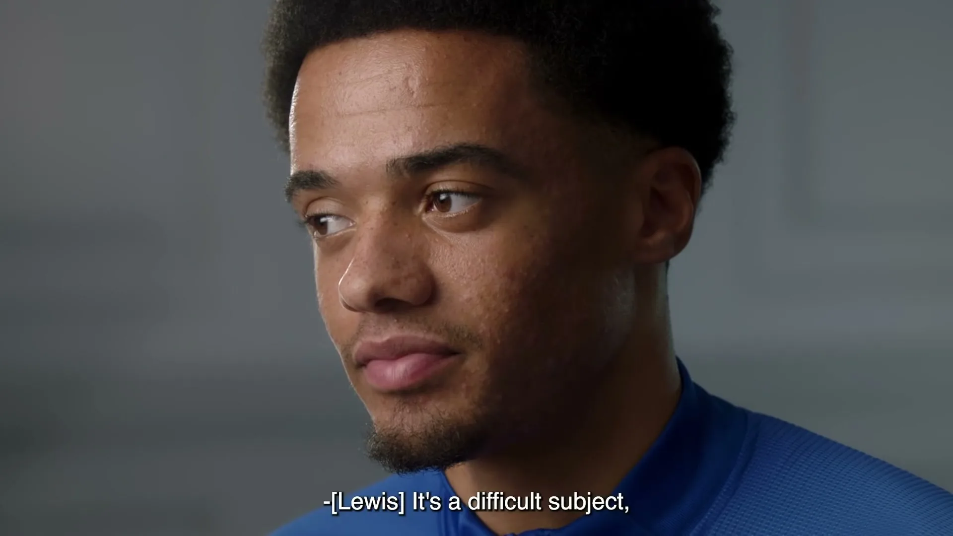 A young Black man with short curly hair and a beard, wearing a blue athletic shirt, looks thoughtfully to the side in a studio setting.