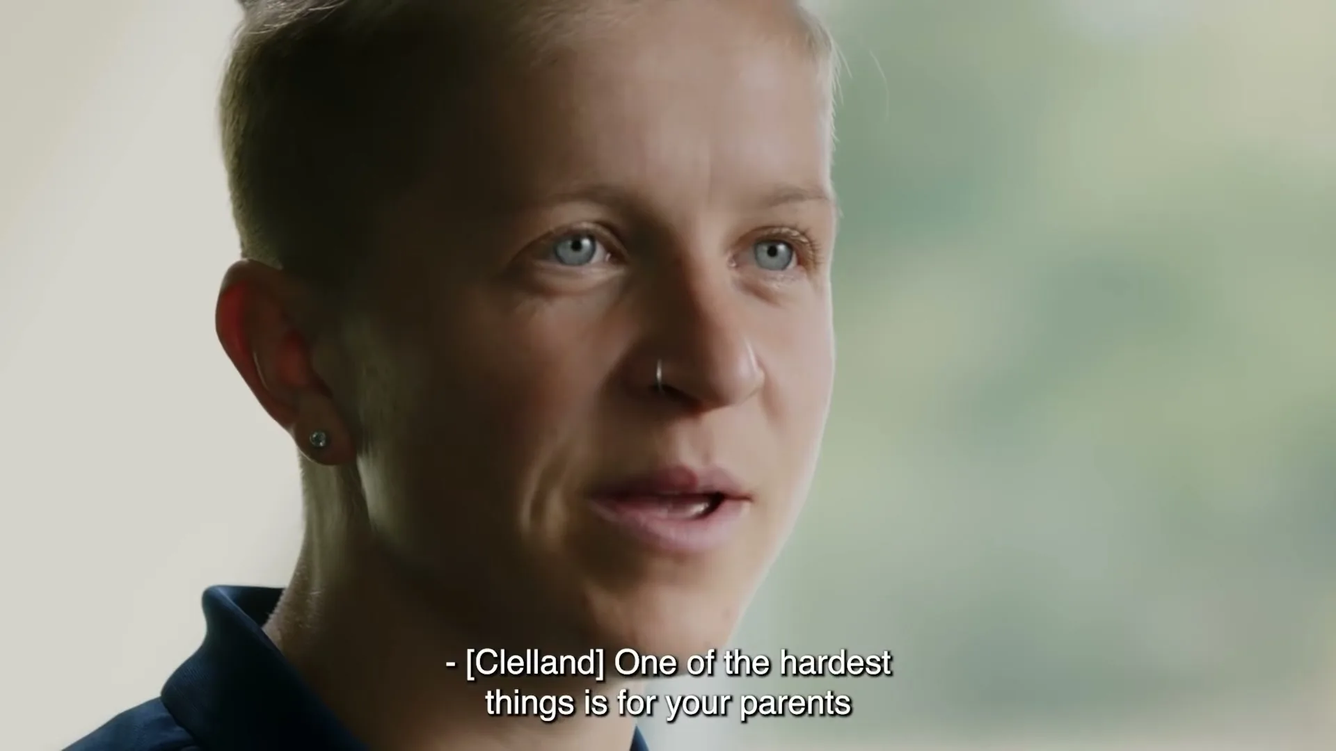 A person with short blonde hair and blue eyes speaks, wearing a dark blue shirt, with a blurred green background.