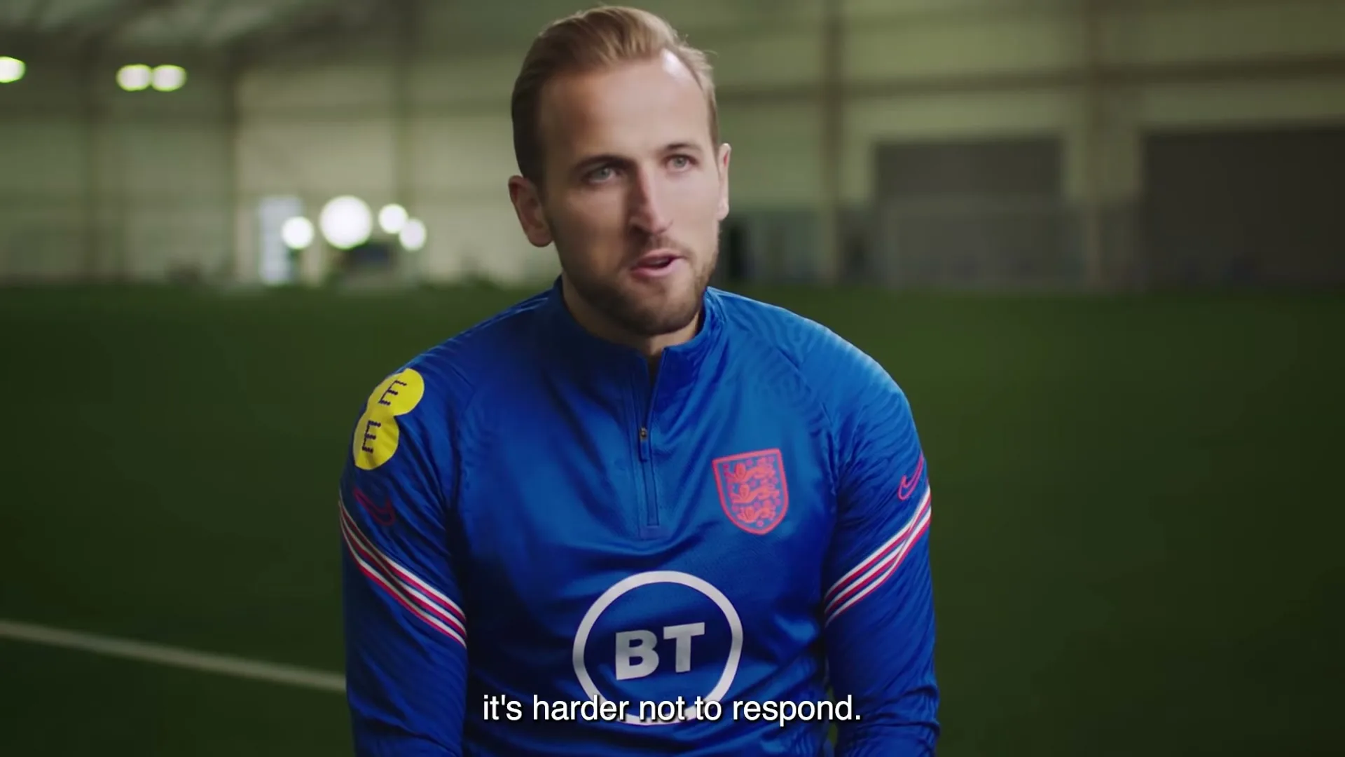 Harry Kane, a male soccer player, speaks directly to the camera while sitting on an indoor soccer field.