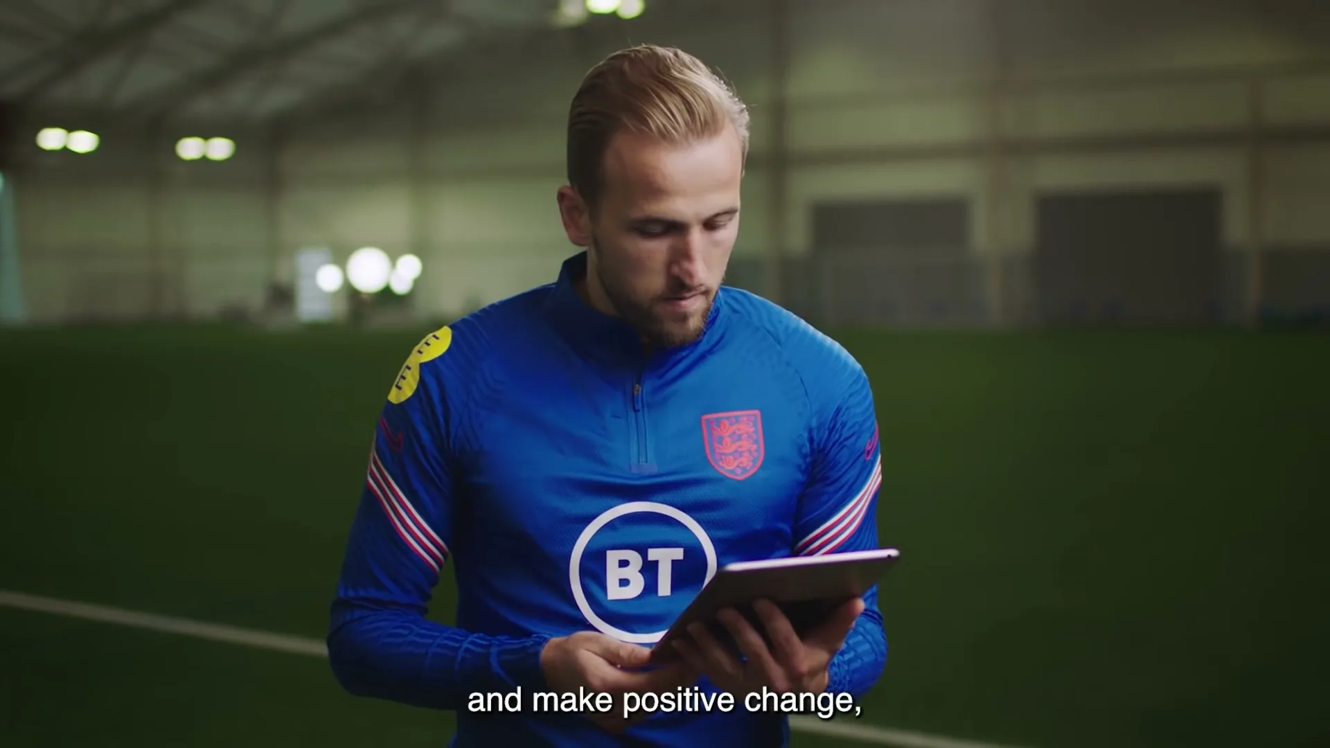 A male footballer in a blue training top looks down at a tablet he is holding in an indoor football pitch.