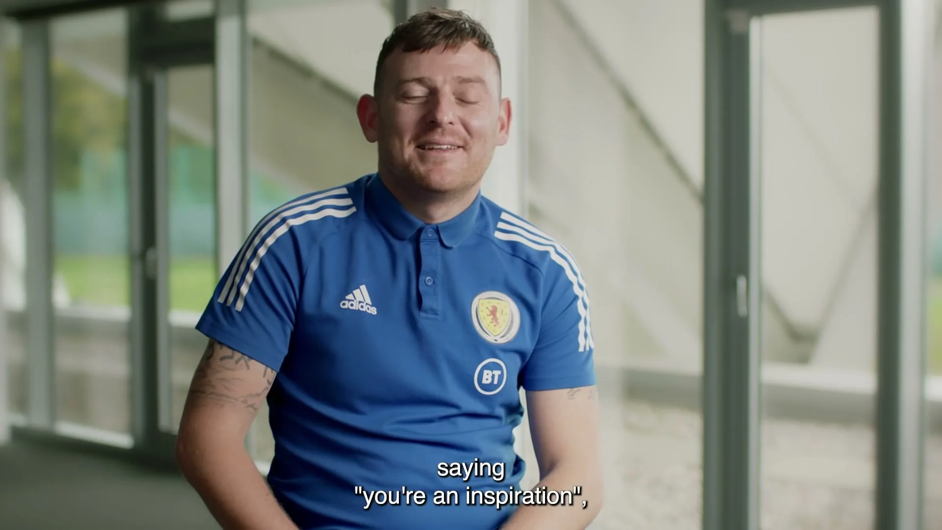 A man in his 20s, wearing a blue polo shirt with a Scottish football crest, smiles at the camera in a modern, bright interior space.