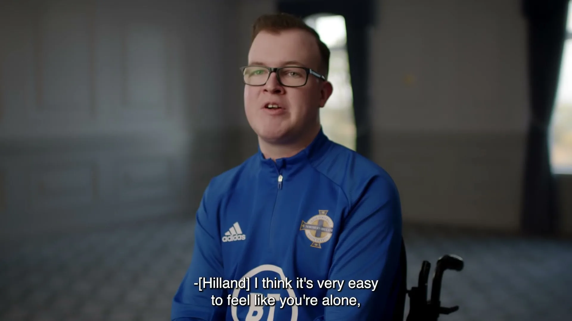 A young man in his 20s, wearing glasses and a blue sports jersey, sits in a wheelchair and speaks directly to the camera in an interior setting.