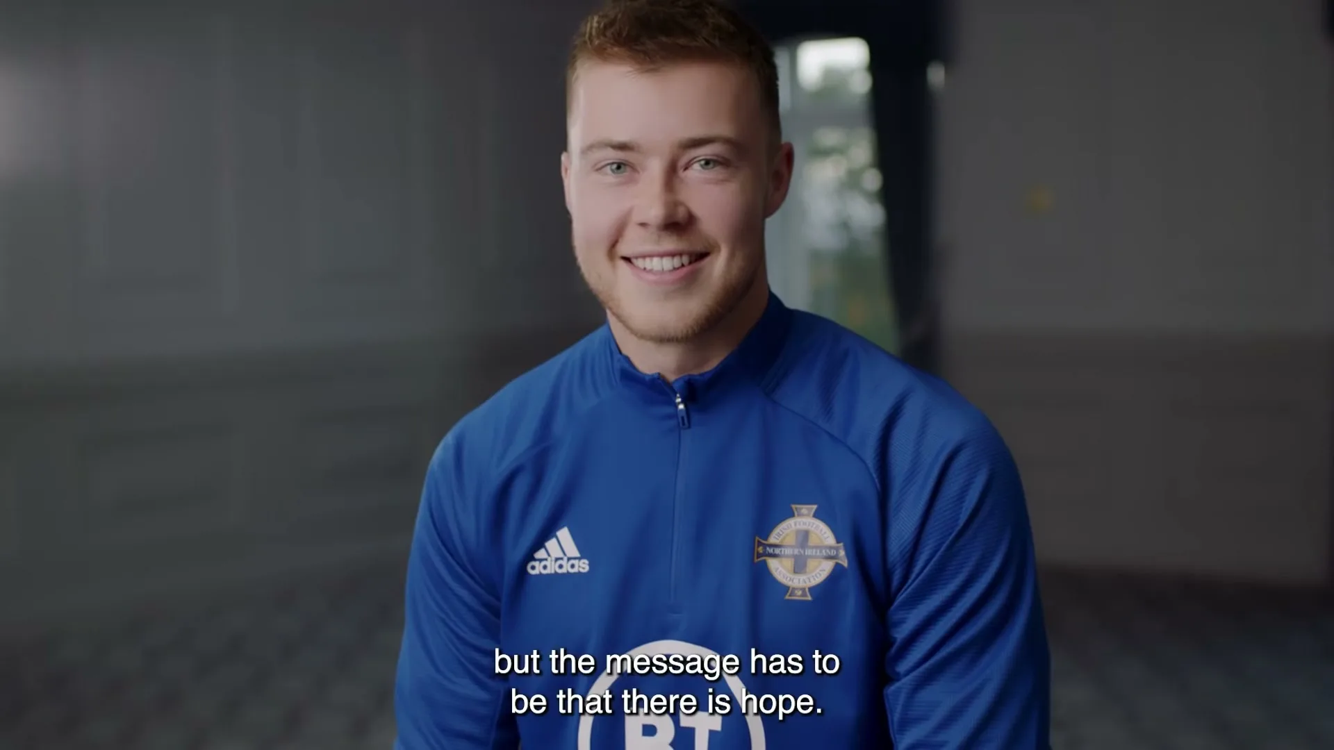 A young male footballer in a blue jersey smiles at the camera, with a blurred background of an indoor space.
