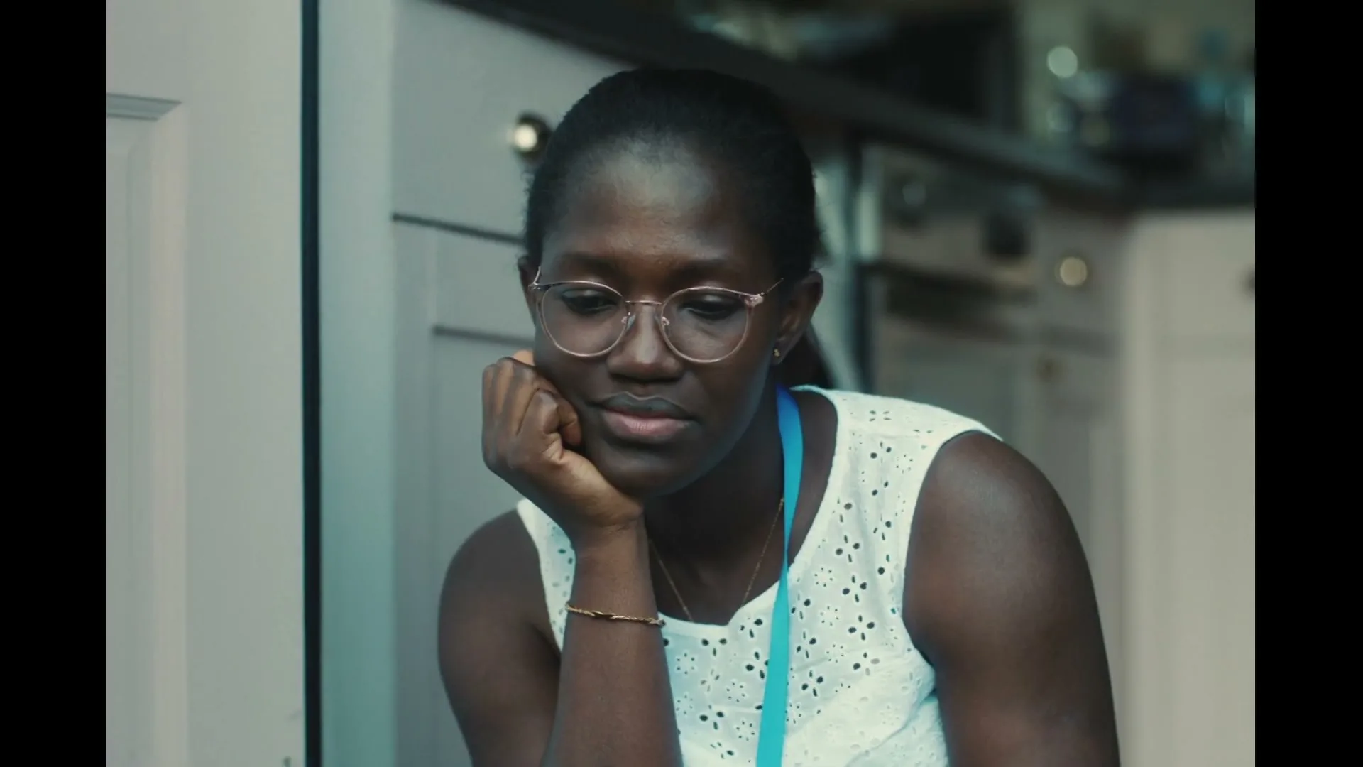 A young Black woman with glasses and a white top sits with her chin resting on her hand, looking down thoughtfully in a kitchen.