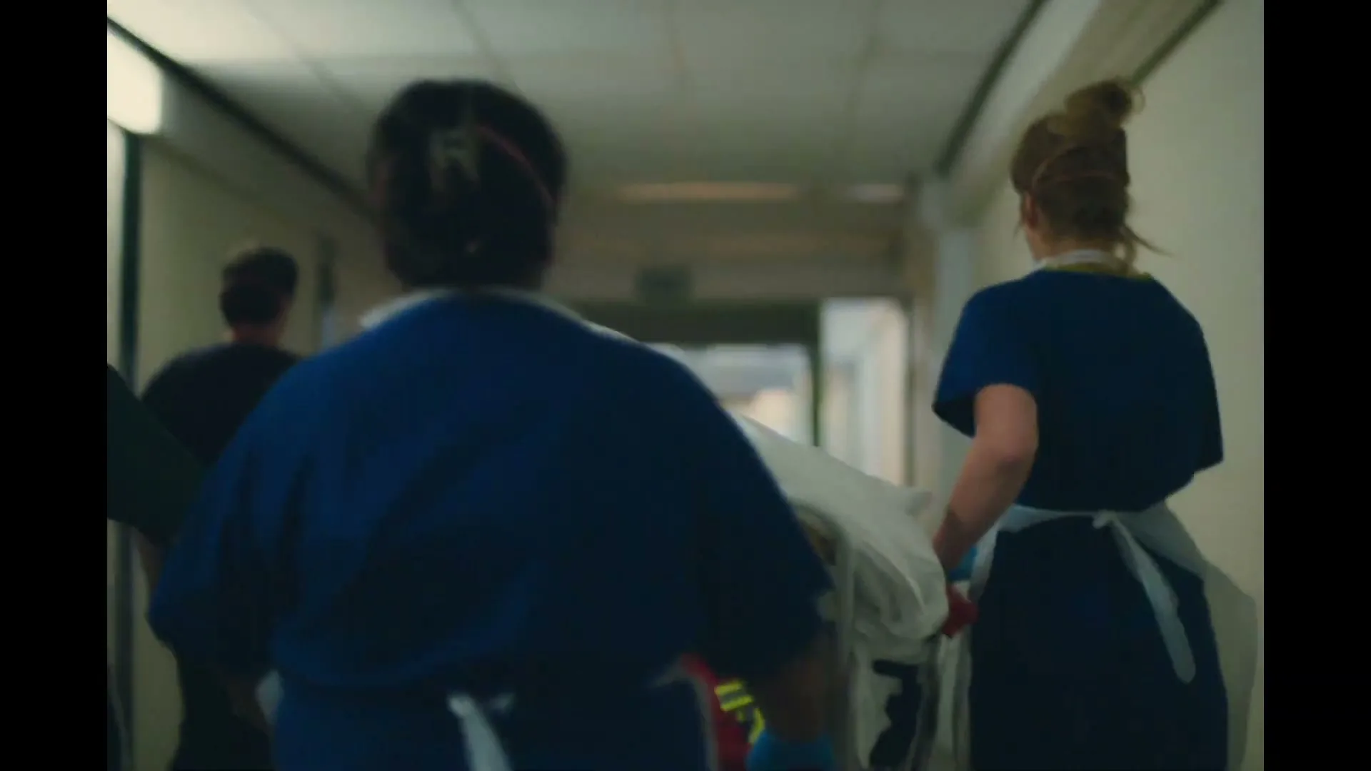 Two medical professionals in blue scrubs and white aprons push a gurney down a hospital hallway, seen from behind.