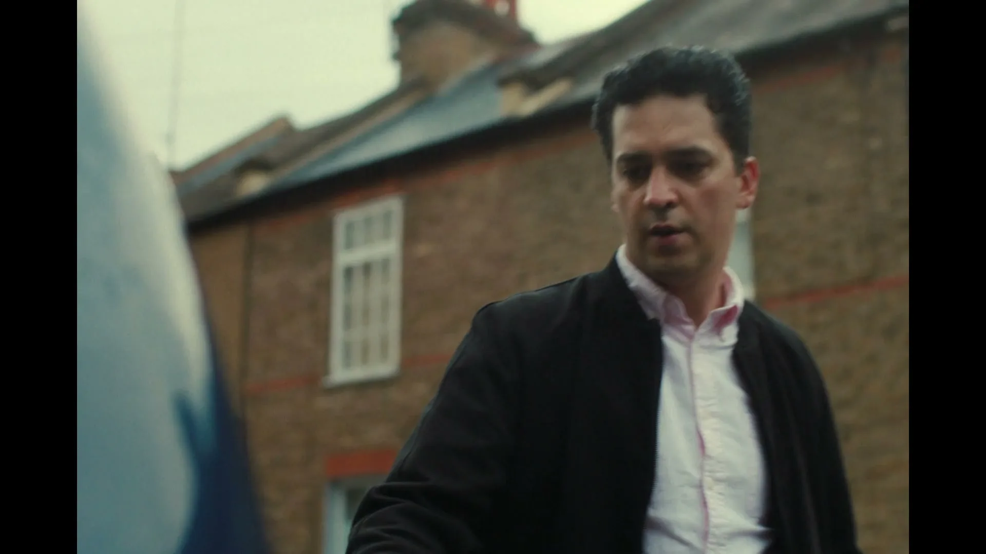 A man in his 30s, wearing a black jacket and pink shirt, looks down and then up, standing next to a car on a residential street.