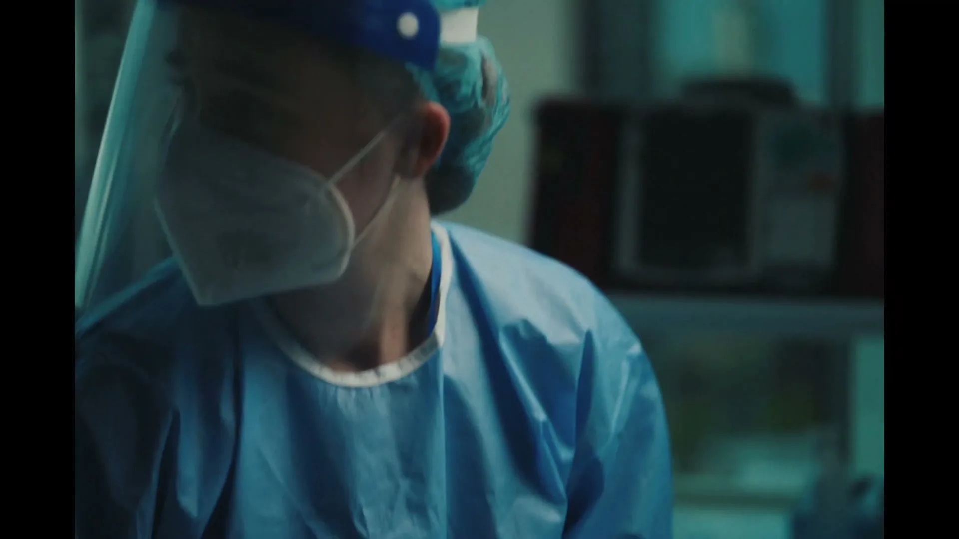 A medical professional in blue scrubs, gloves, face mask, and shield holds the hand of a patient in a hospital bed, then looks down sadly.