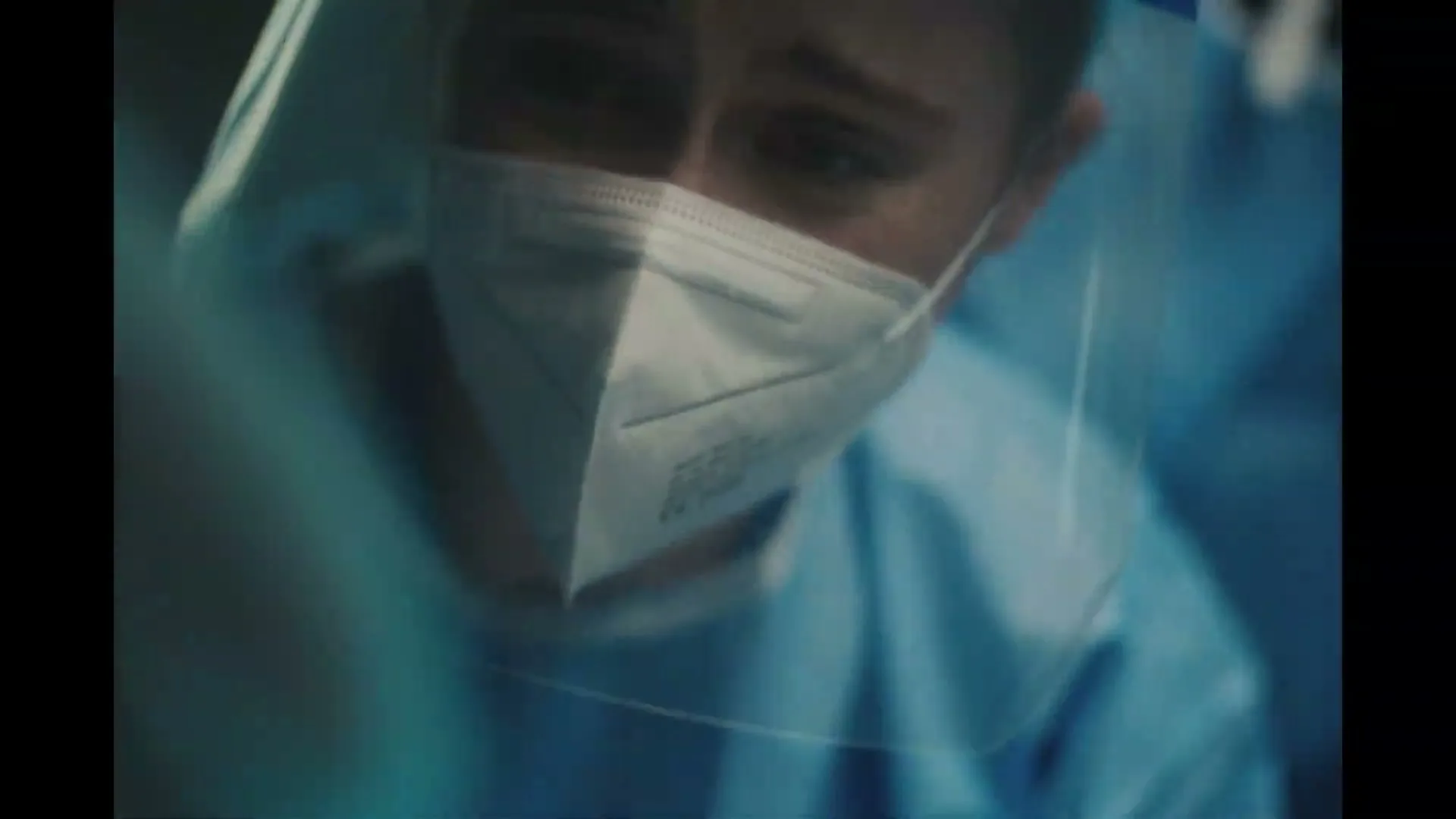A healthcare worker in scrubs, face mask, and face shield looks down with a somber expression in an interior medical setting.