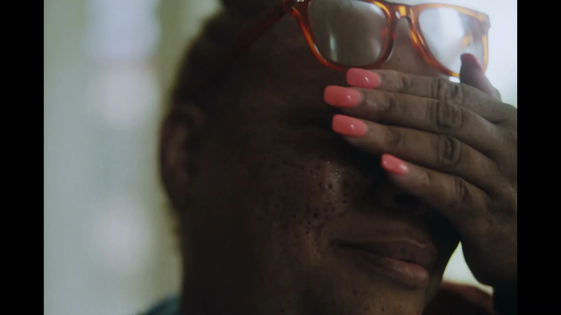 A woman with orange glasses and freckles cries, covering her face with her hand, in a soft-lit interior.