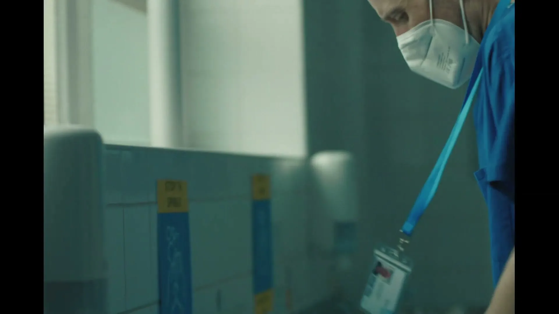 A bald male healthcare worker in blue scrubs and a white mask washes his hands at a sink in a hospital bathroom.