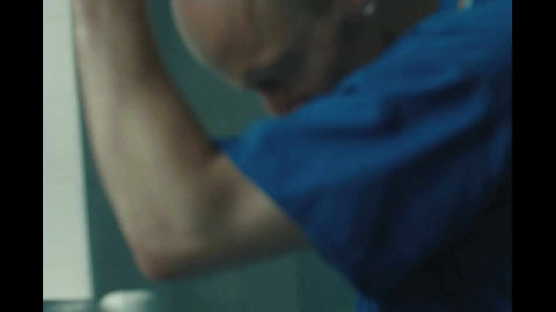 A bald male healthcare worker in blue scrubs removes his face mask, looking down with a pained expression in a hospital room.