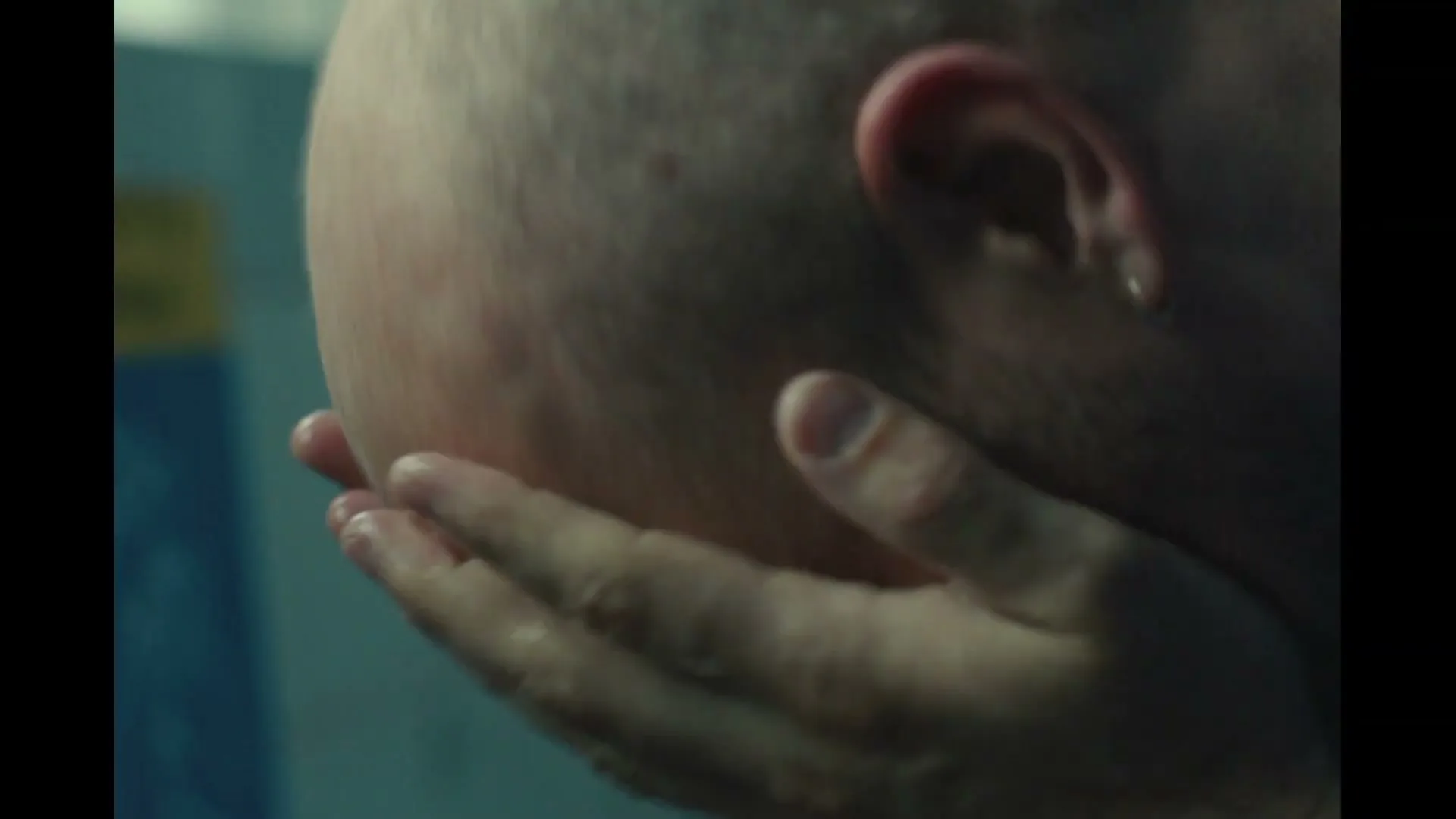 A male healthcare worker in blue scrubs is seen from behind, washing his face with water in a bathroom, looking distressed.