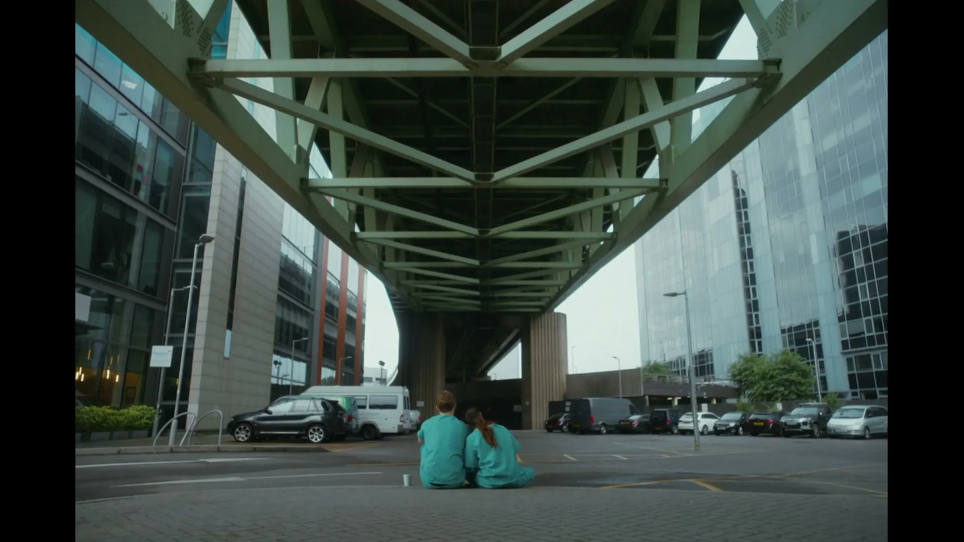 Two medical professionals in scrubs sit on the ground under a large bridge, surrounded by modern buildings and parked cars.