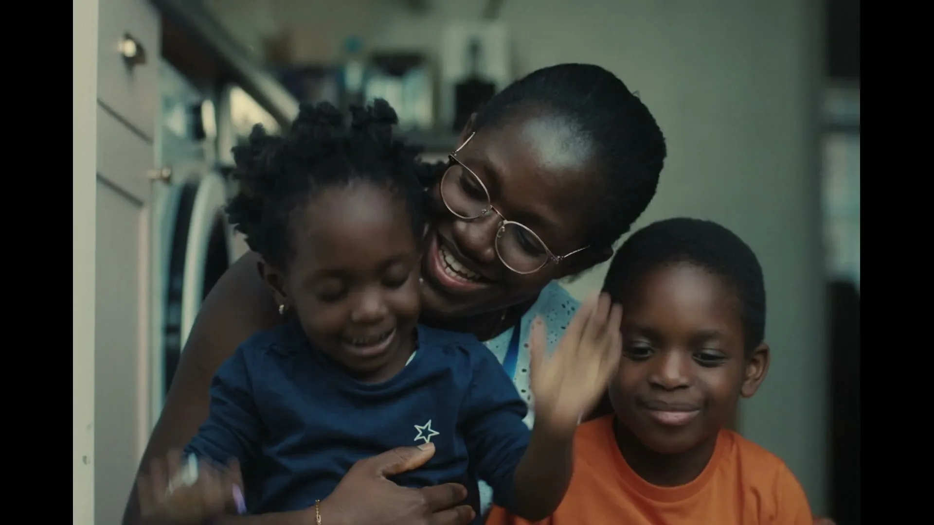 A Black mother with glasses smiles while holding her two young children, a girl and a boy, who are also smiling and playing, in a domestic setting.