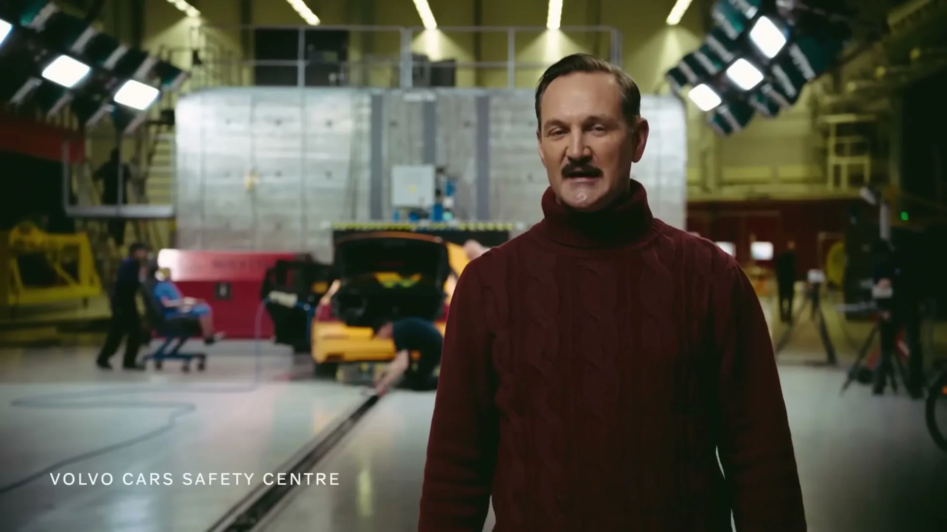 A man with a mustache and a red turtleneck sweater stands in a car crash test facility, speaking to the camera while technicians work on a yellow car in the background.