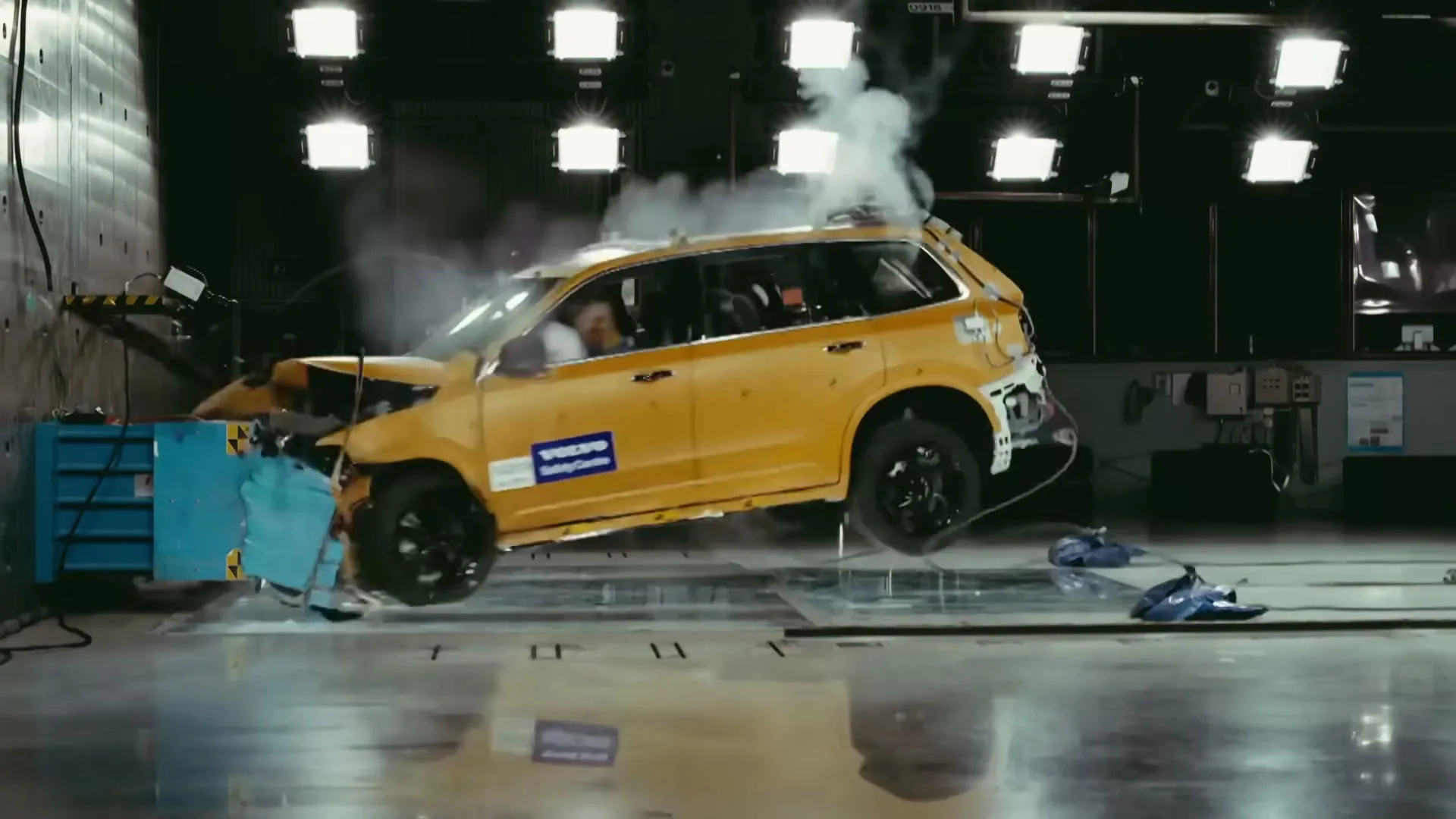 A yellow car crashes into a blue barrier in a crash test facility, with smoke and debris flying as the vehicle crumples on impact.