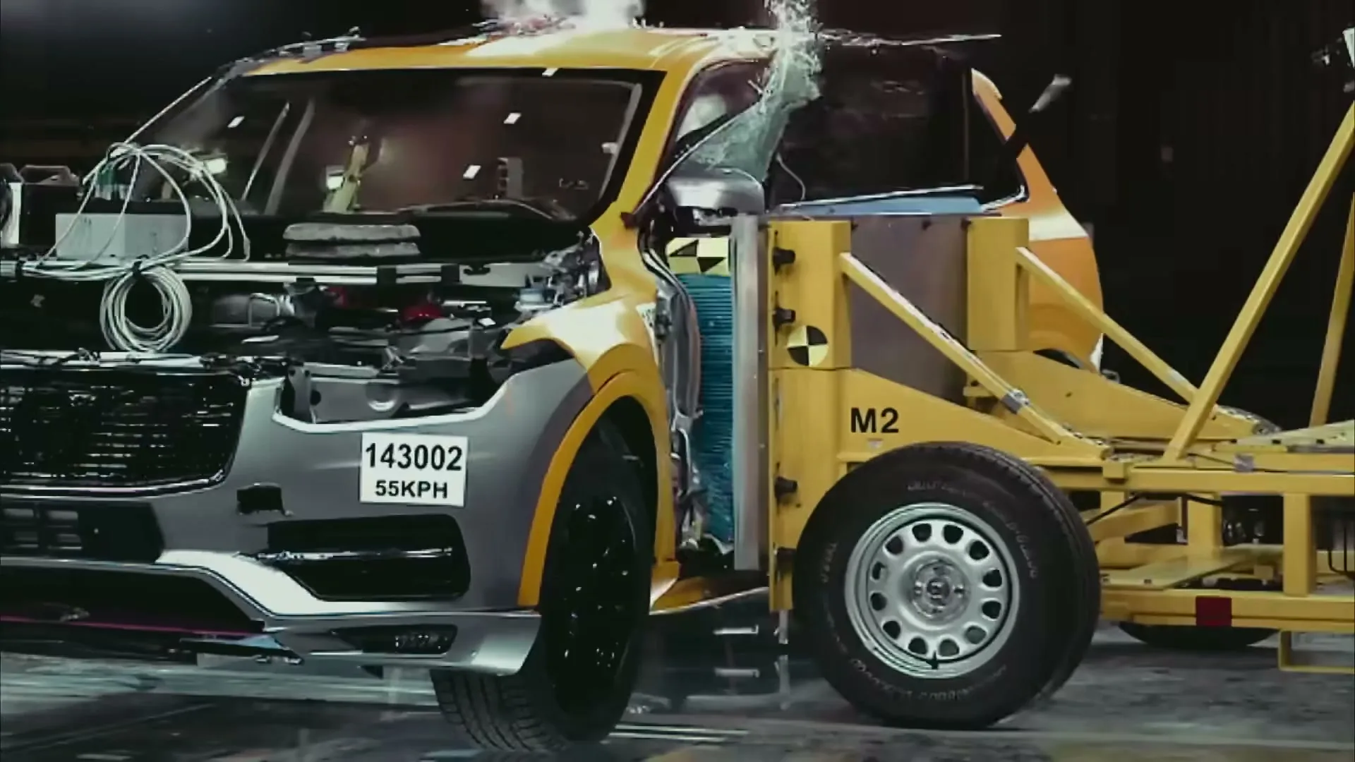 A yellow and grey Volvo SUV undergoes a side-impact crash test, with the side of the car crumpling and glass shattering in a controlled environment.