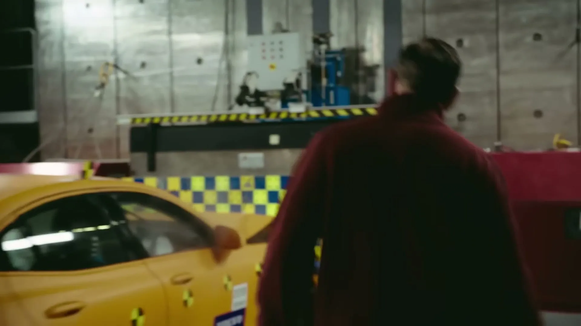 A man in a red sweater walks past a yellow crash-test car in a brightly lit industrial crash-test facility.