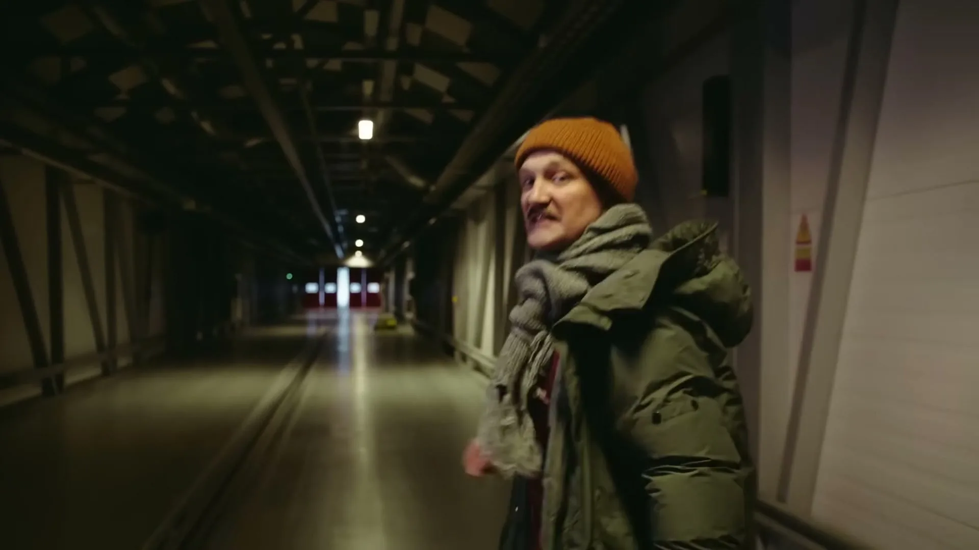 A man in a winter coat and hat walks backward down a long, dark industrial hallway, looking over his shoulder at the camera with a mischievous expression.