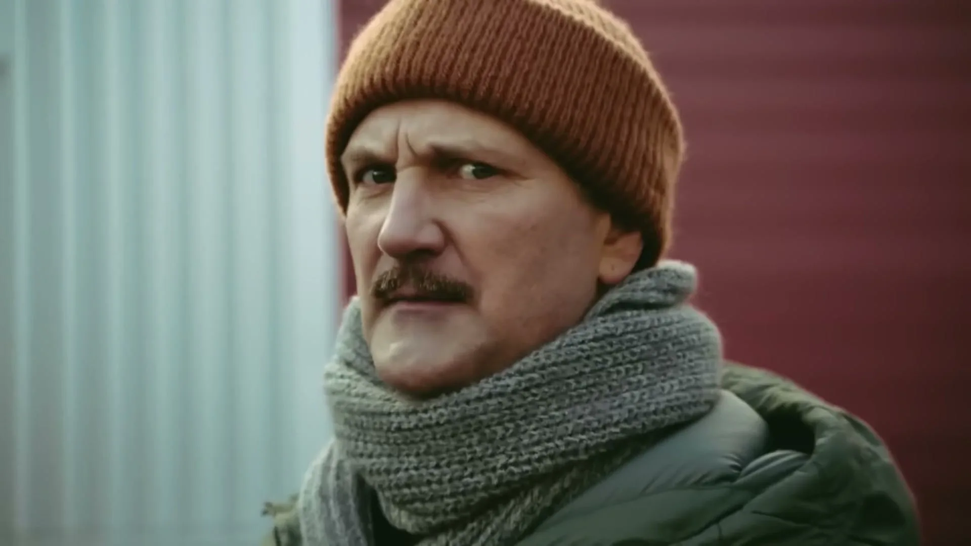 A middle-aged man with a mustache, wearing a brown beanie and a green jacket, looks surprised and then concerned as he turns his head in an exterior setting.