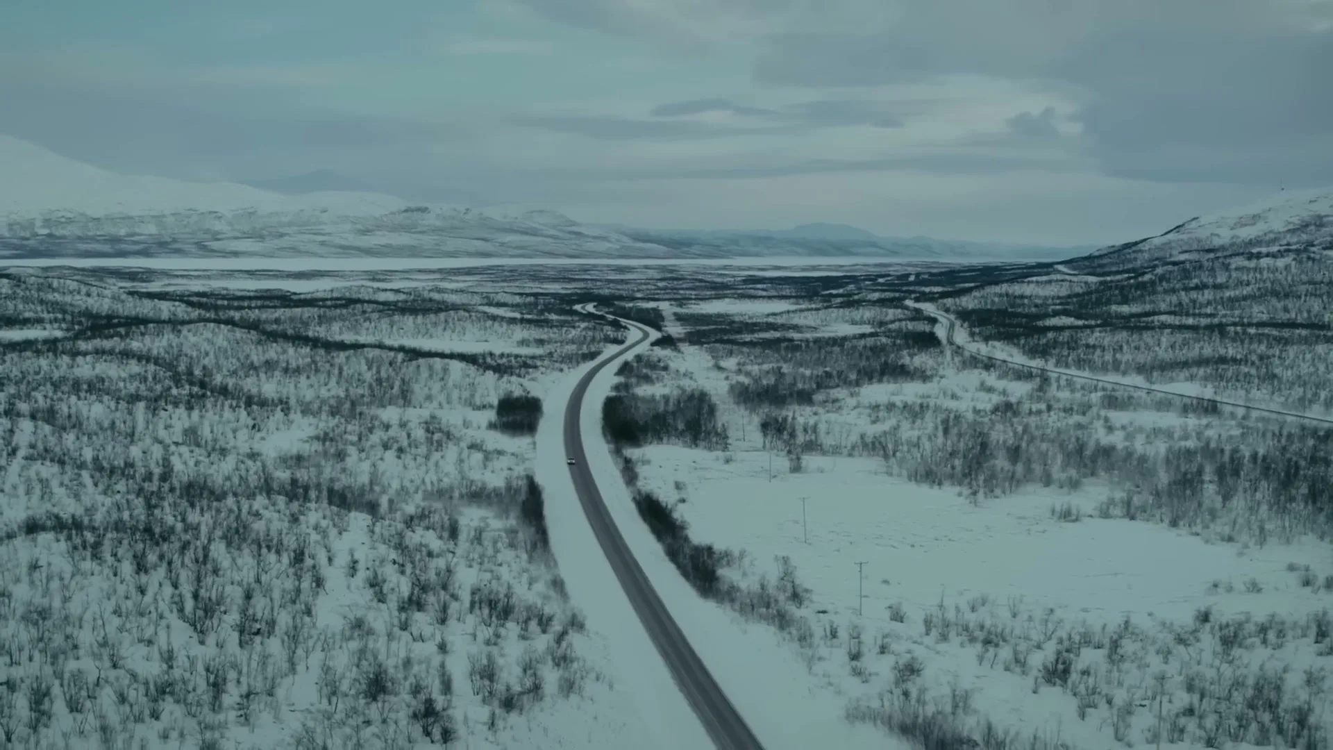 An aerial wide shot shows a white Volvo car driving on a snowy road through a vast, snow-covered forest with mountains in the distance under a cloudy sky.