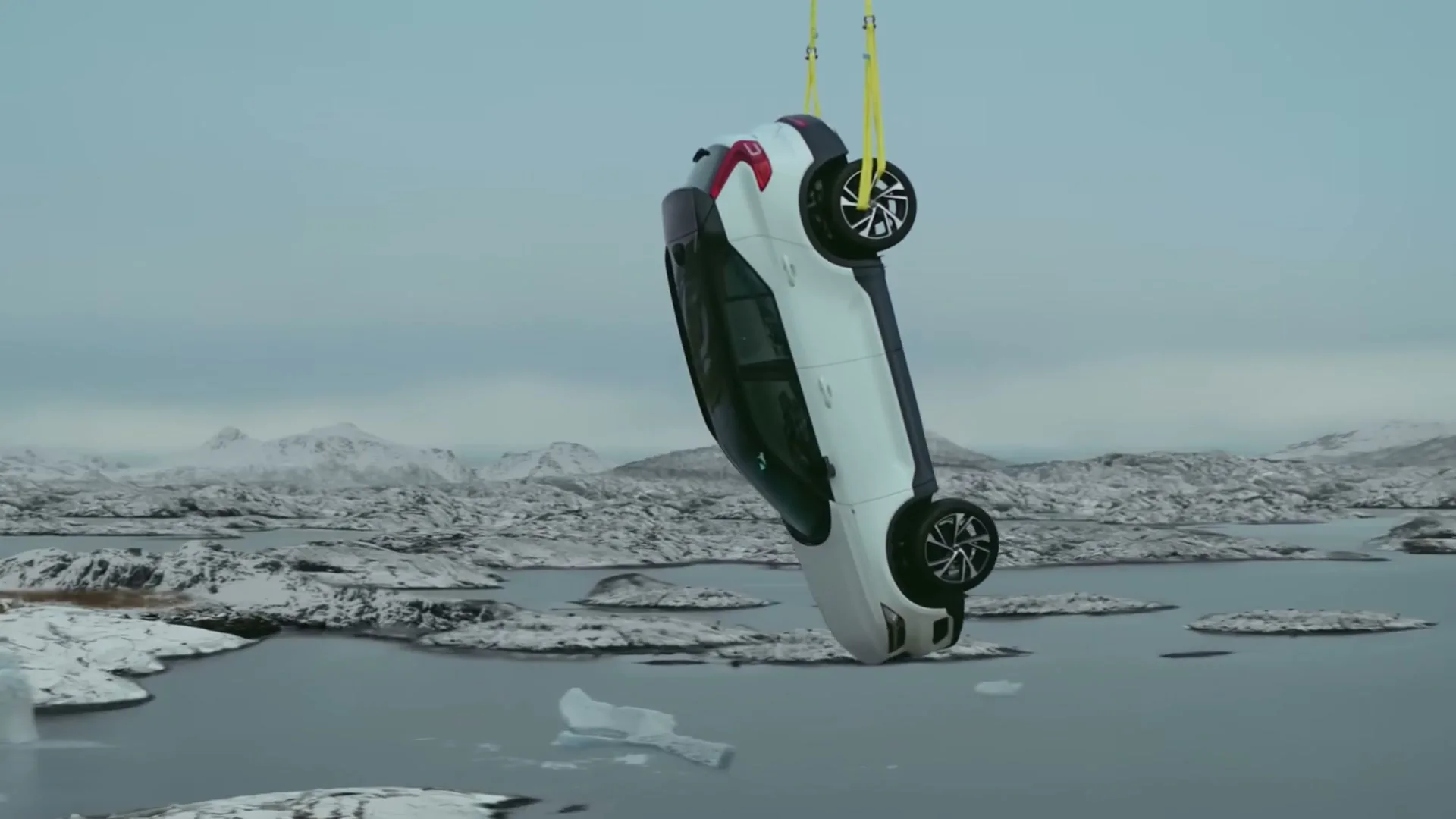 A white Volvo car is suspended upside down by yellow straps over a vast, icy landscape with snow-covered mountains and a dark body of water.