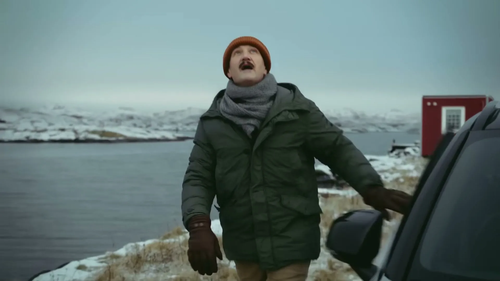 A man with a mustache, wearing a winter coat and hat, stands next to a car on a snowy cliff overlooking the sea, looking up in surprise.