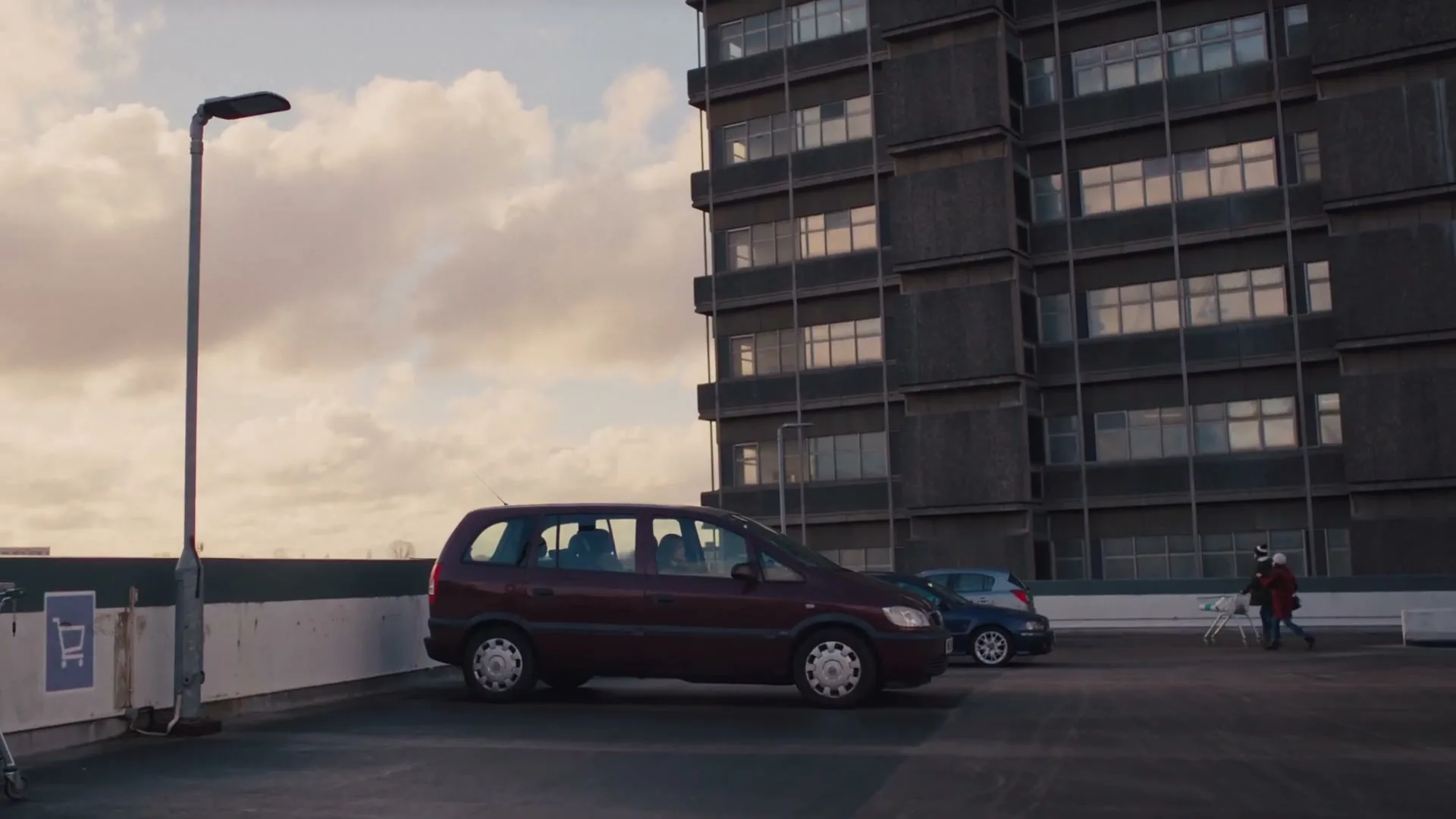 A woman and a child walk across a rooftop parking lot towards a tall, brutalist building under a cloudy sky.