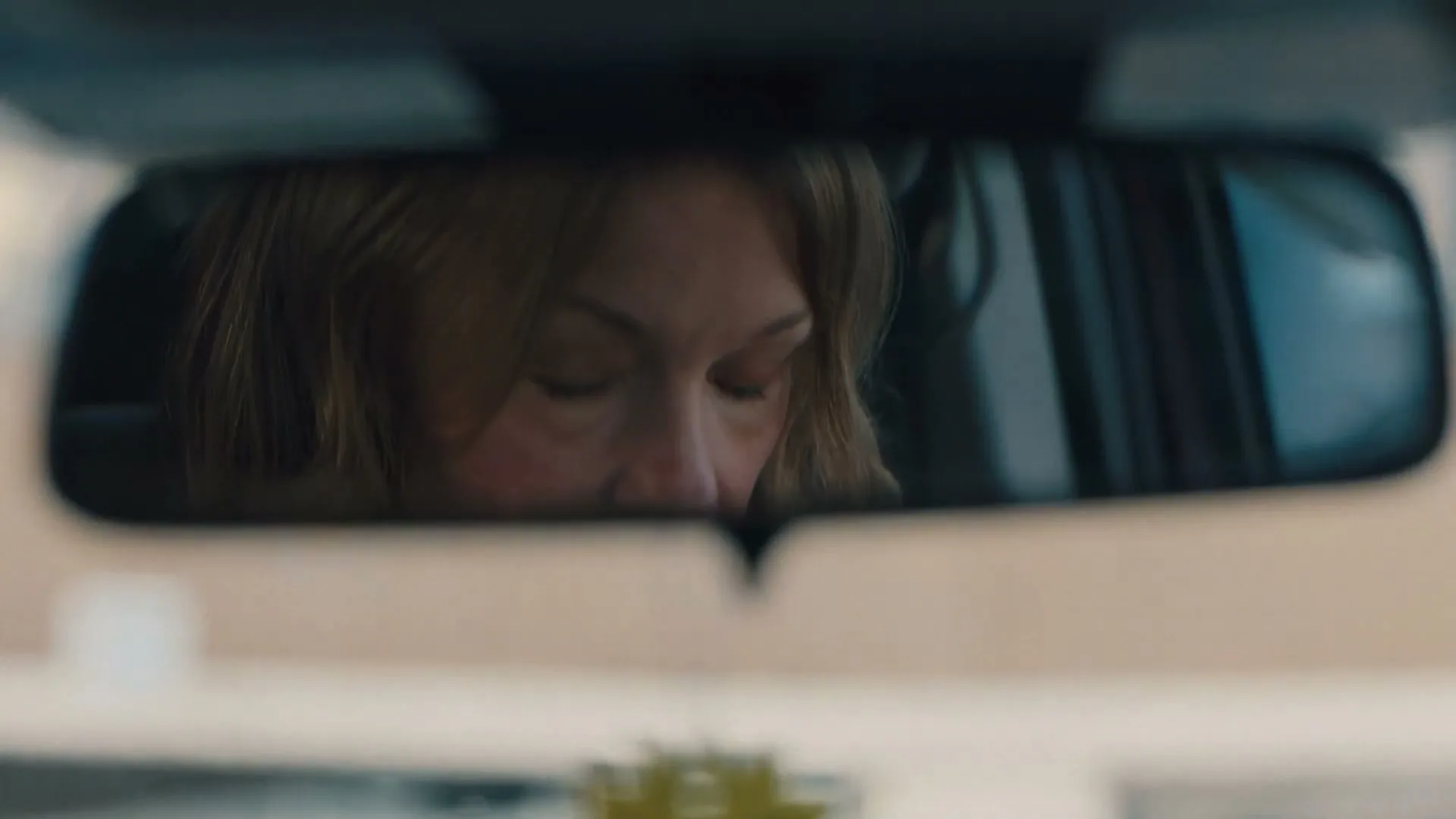 A woman with blonde hair is seen in a car's rearview mirror, looking up with a concerned expression.