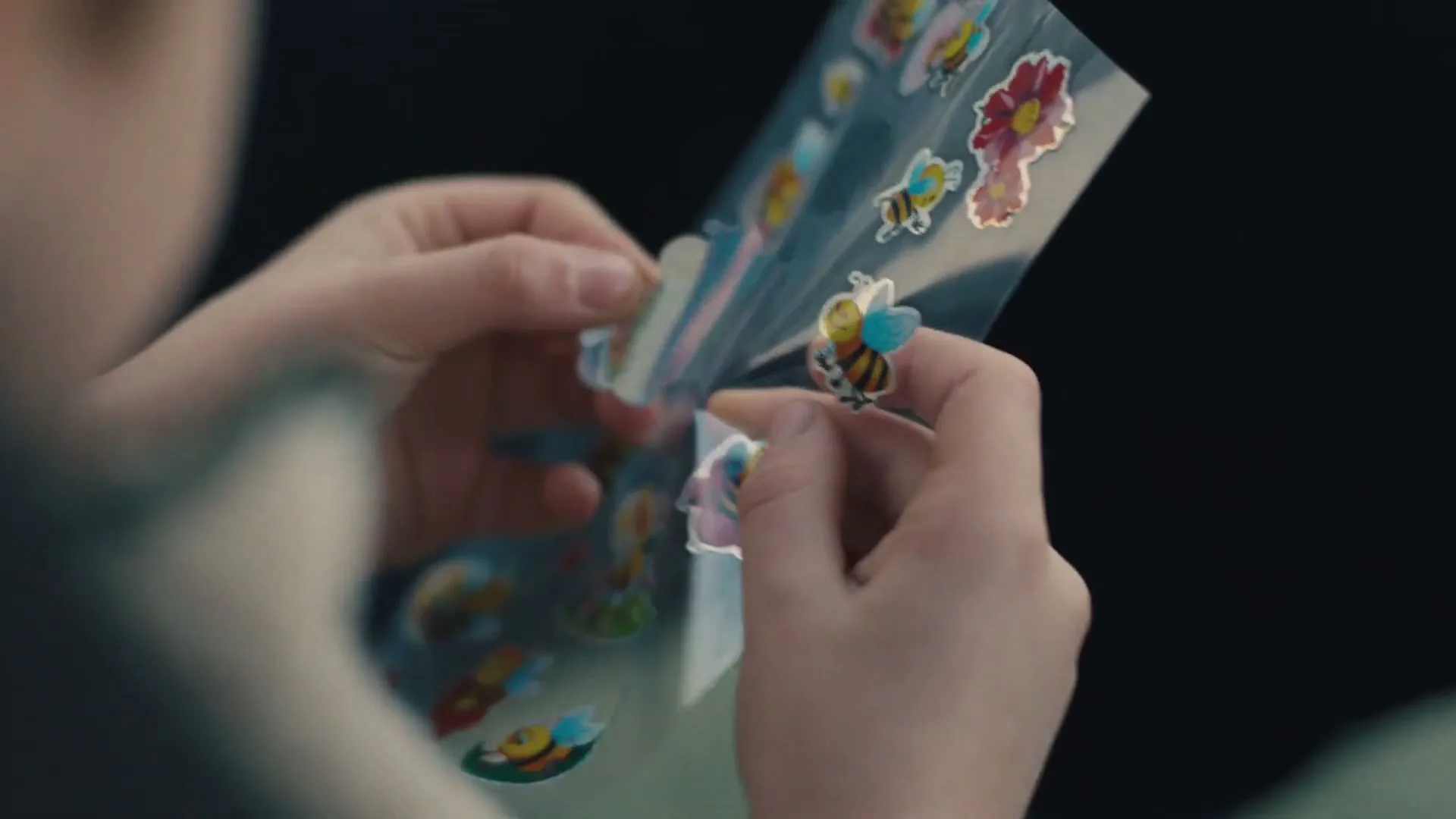 A child's hands carefully peel a bee sticker from a sheet, with a blurred background suggesting an interior setting.