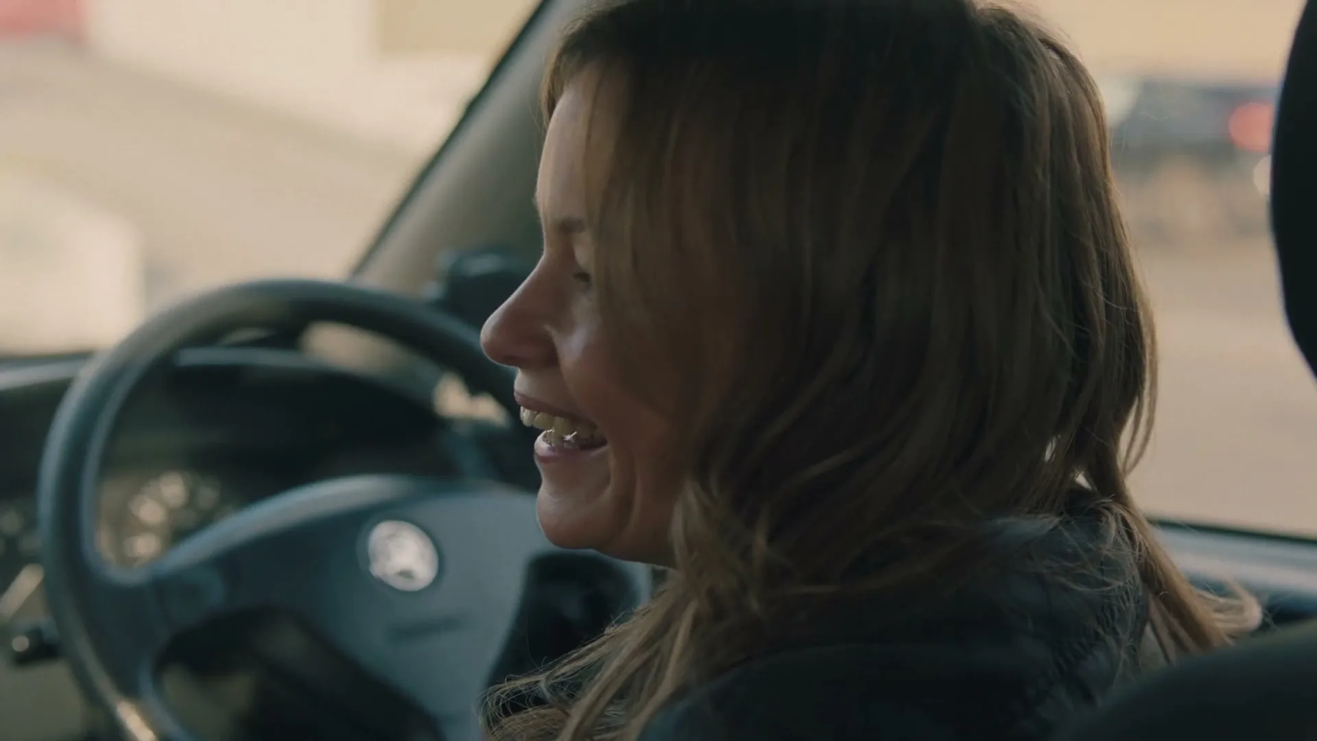 A woman with long brown hair, wearing a dark jacket, smiles and talks while driving a car during the day.