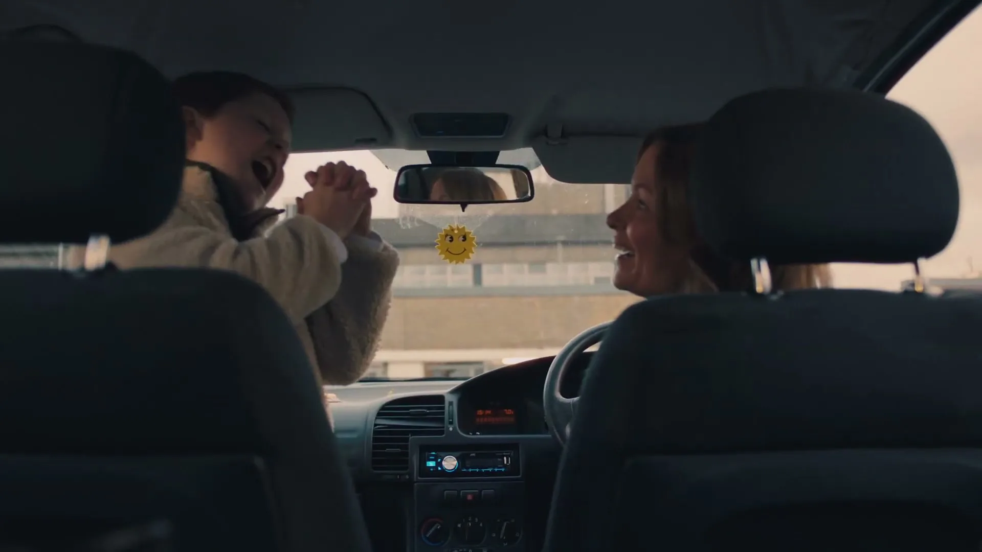 A young boy in a cream jacket claps his hands excitedly while his mother, with brown hair, smiles at him from the driver's seat of a car.