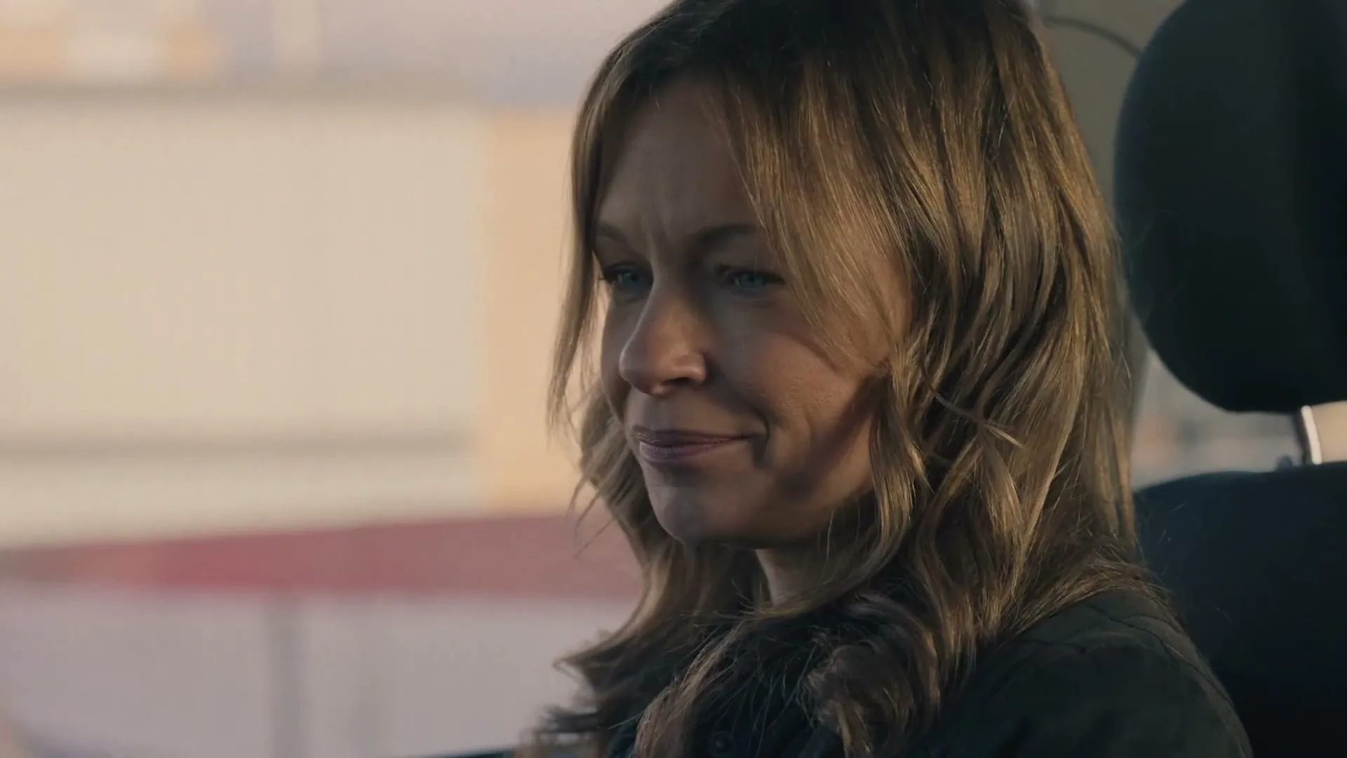 A woman with long brown hair sits in a car, looking down and then up with a slight smile, as if listening to someone.
