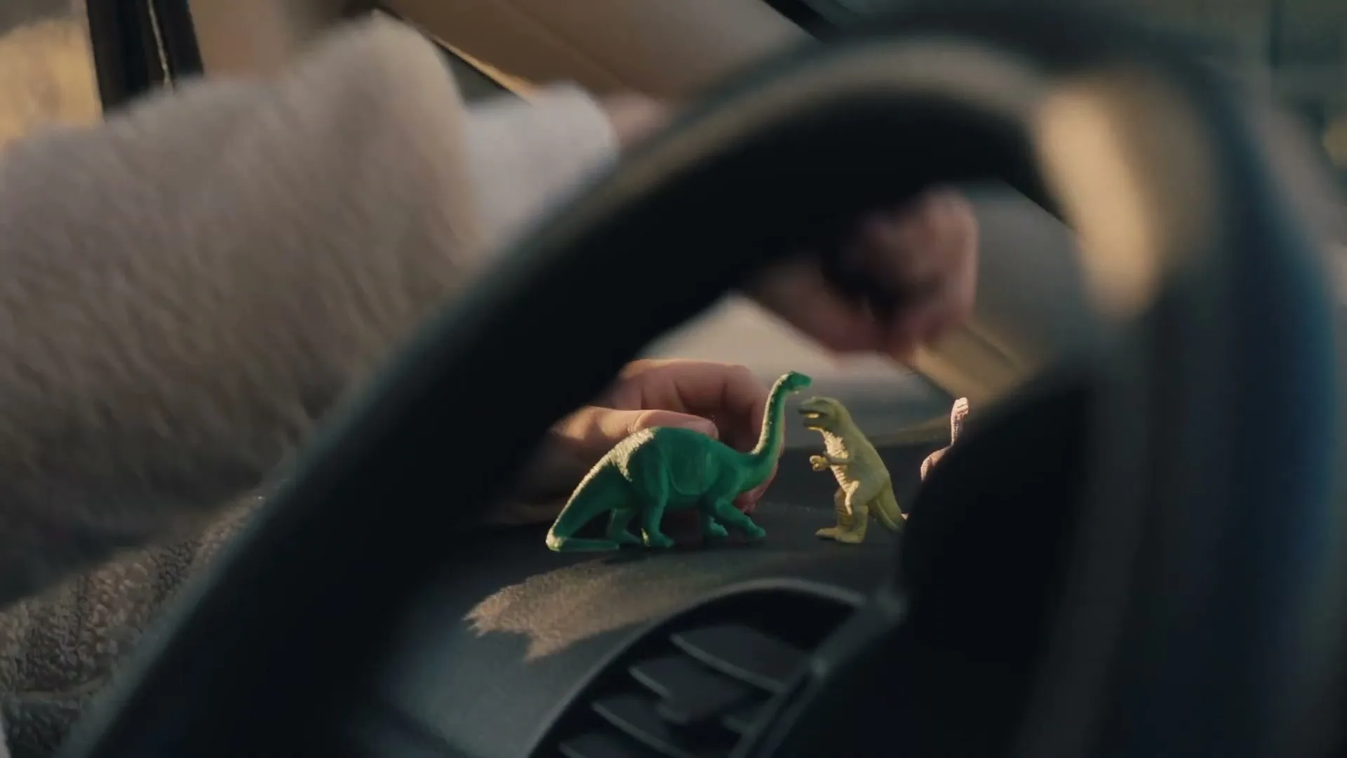 A child's hands play with two toy dinosaurs on a car dashboard, seen through the steering wheel, in a car interior.
