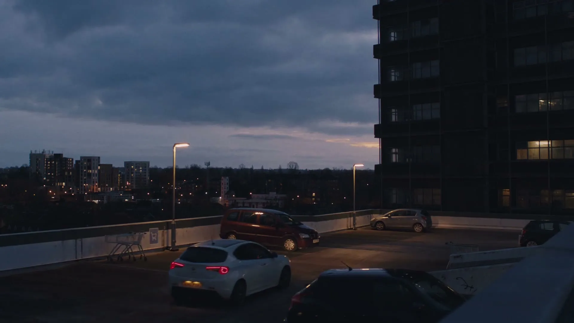 A wide shot shows a dark, multi-story car park rooftop at dusk, with several cars parked and city lights visible in the background.