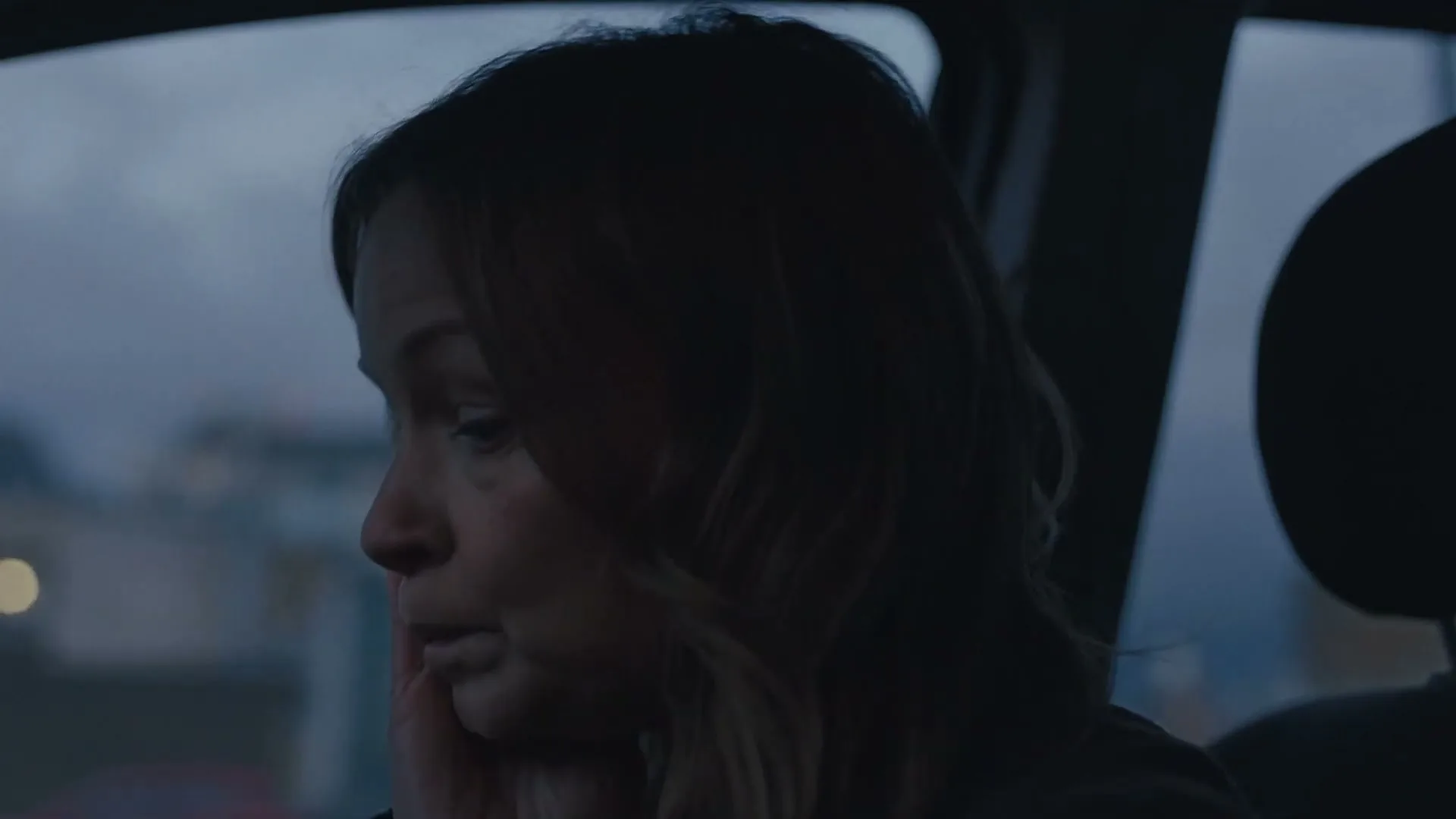 A woman with light brown hair sits in a car, looking out the window with a somber expression as she whispers to herself.