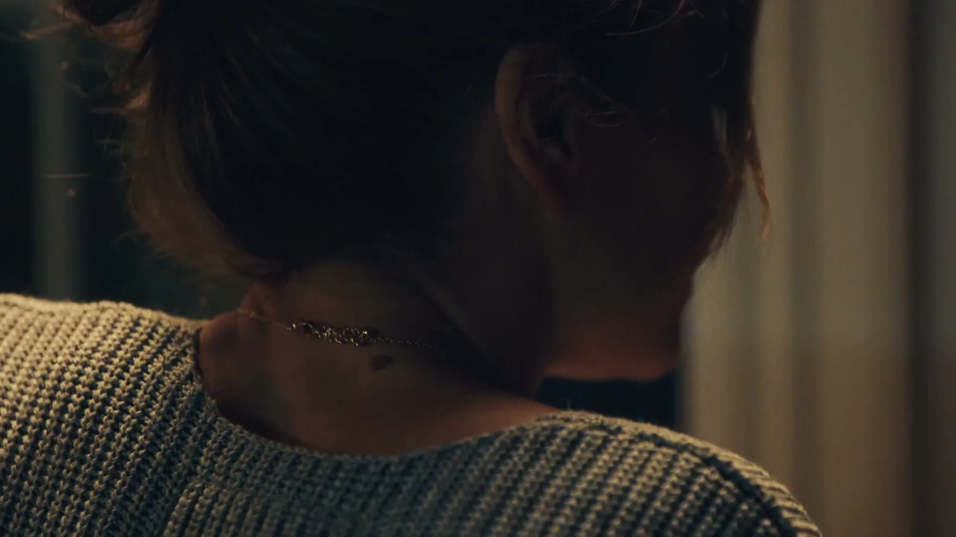 A woman with her hair tied back, wearing a grey knitted sweater and a delicate necklace, turns her head to the right in a dimly lit interior.