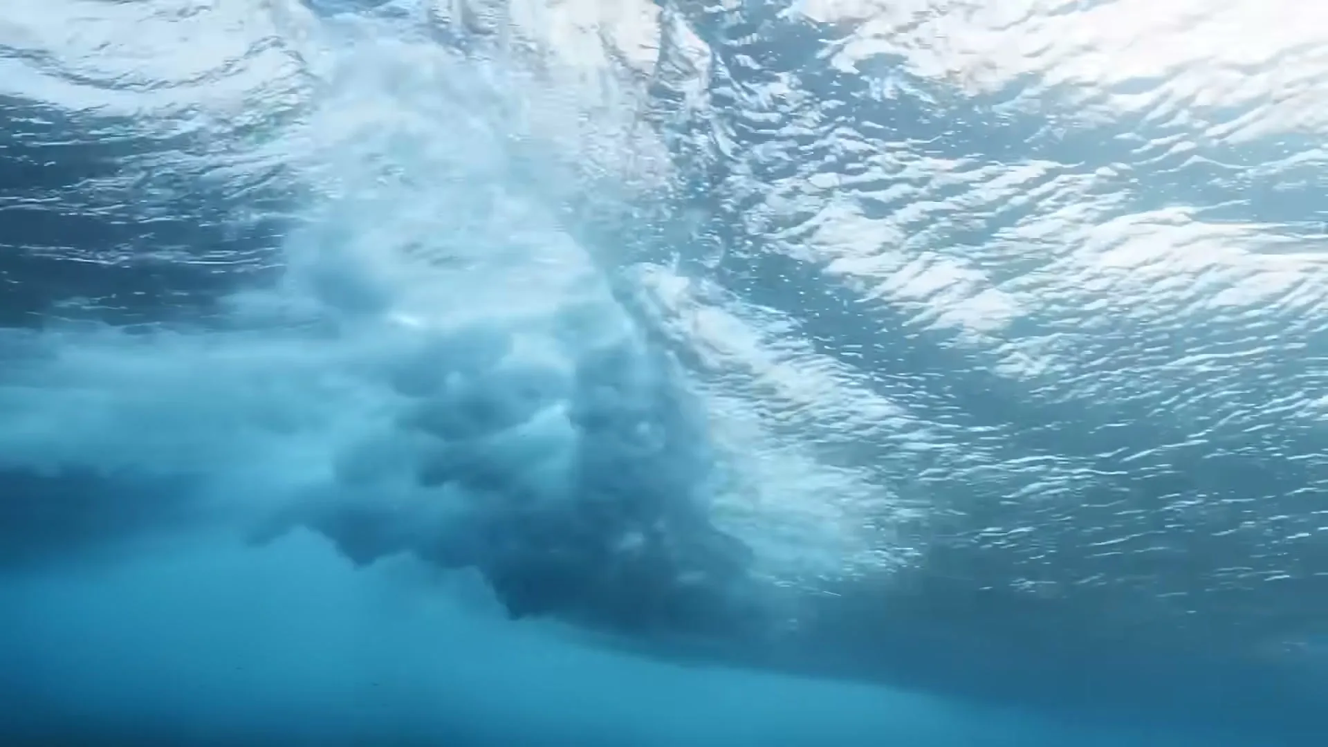 An underwater shot shows the foamy underside of a breaking wave, with sunlight dappling through the surface.