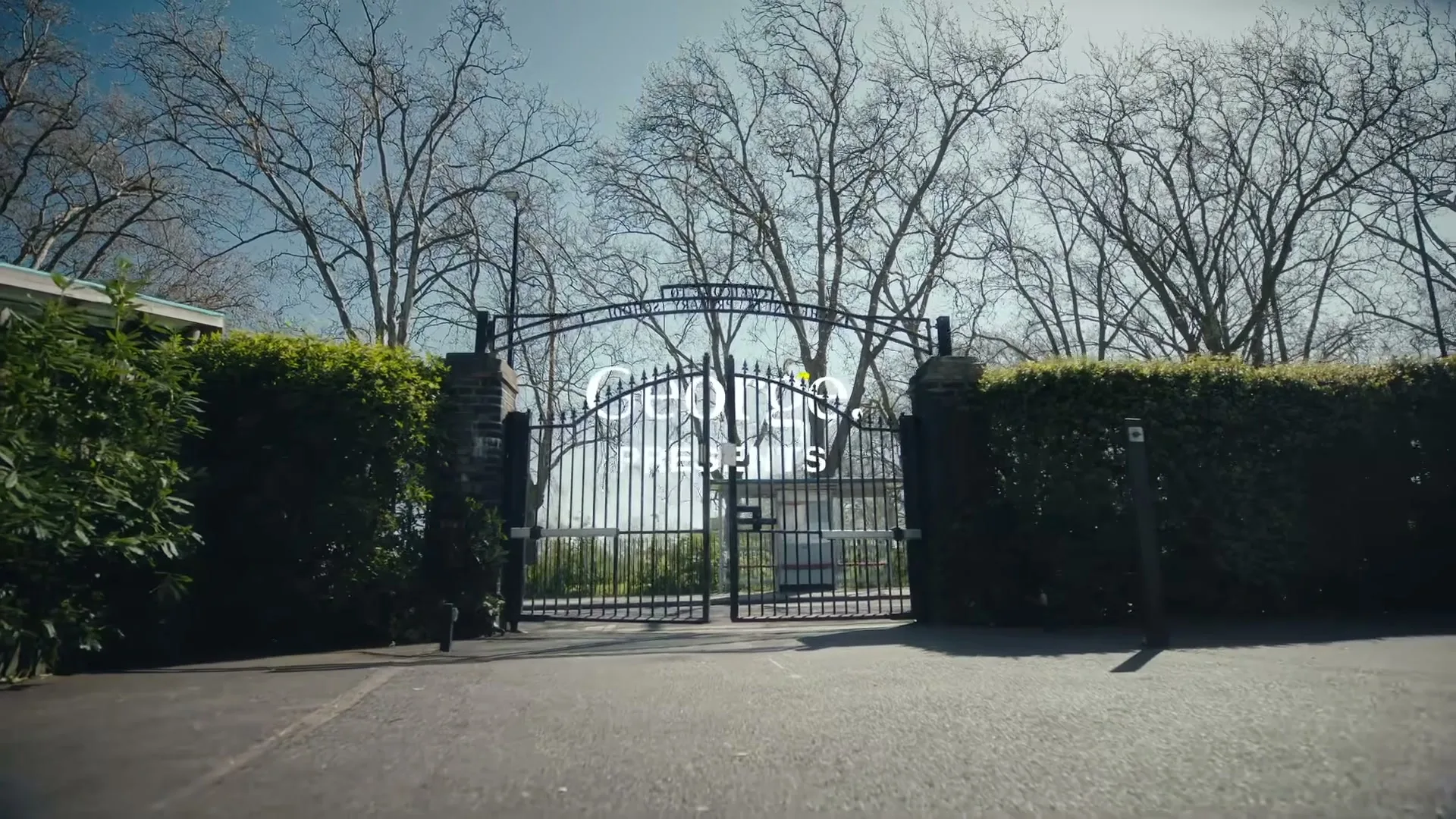 A wide shot of a large, ornate black metal gate with "George at ASDA" text superimposed, surrounded by green hedges and bare trees under a bright sky.